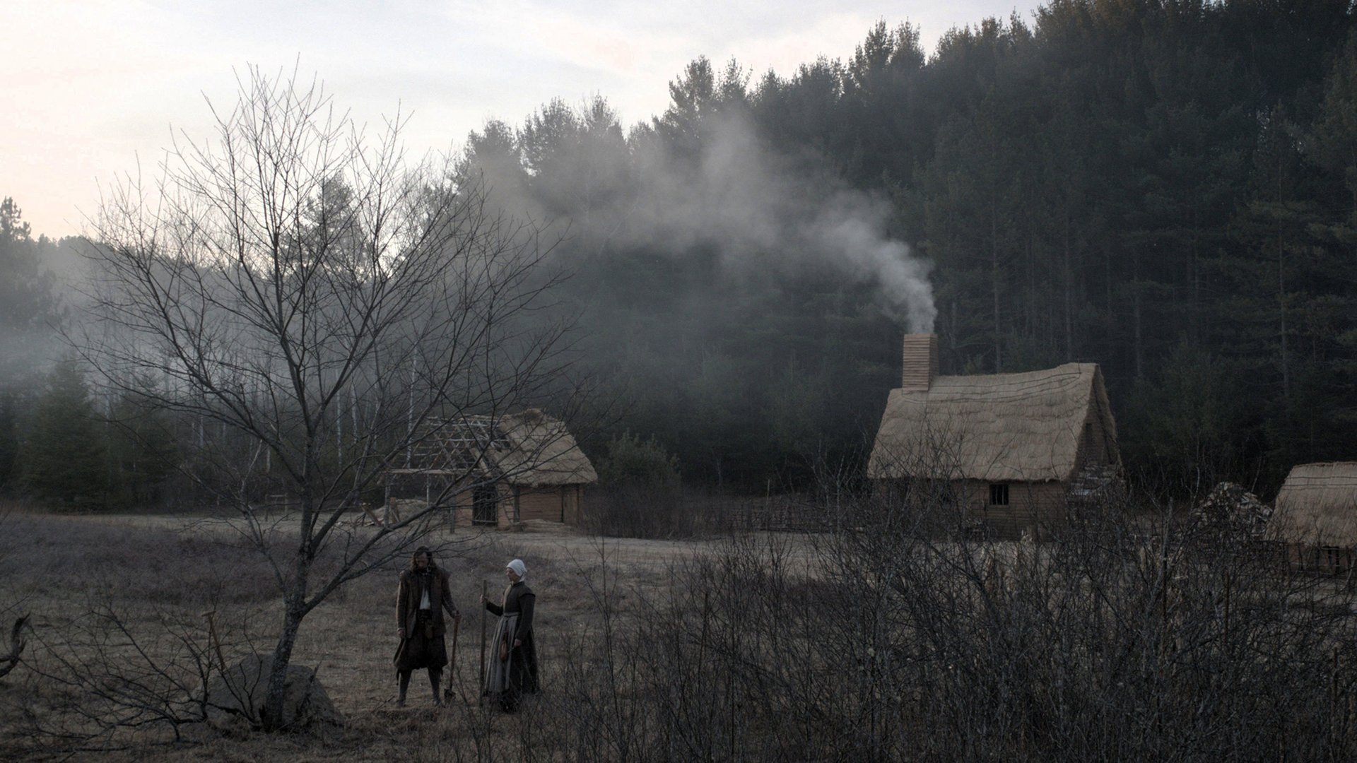 Czarownica Bajka ludowa z Nowej Anglii The Witch kadr | film.org.pl Kadr z filmu Czarownica: Bajka ludowa z Nowej Anglii: dwie postacie w ciemnych strojach idą przez zamglony pusty teren obok drewnianych chat krytych strzechą, z których unosi się dym na tle lasu. folk horror