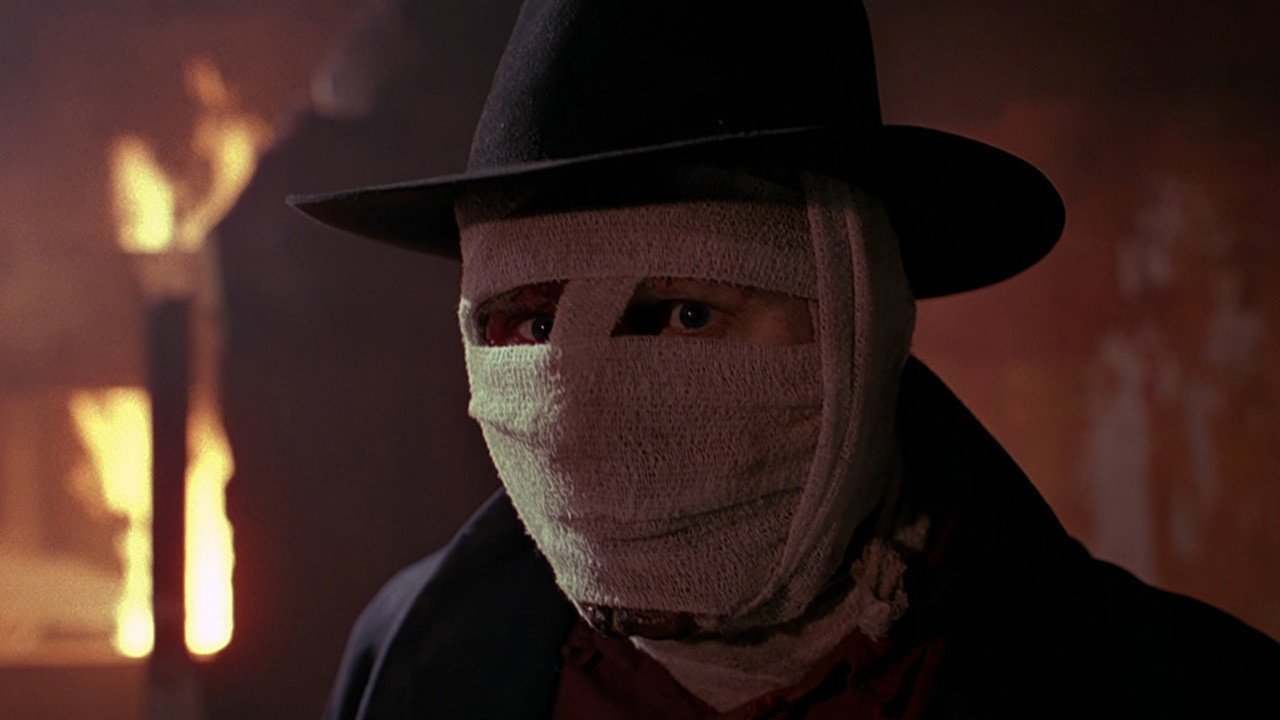 Człowiek Ciemności Darkman kadr | film.org.pl Kadr z filmu Człowiek ciemności: mężczyzna w czarnym kapeluszu i owinięty bandażami twarz, w tle płomienie. superbohaterskie horrory
