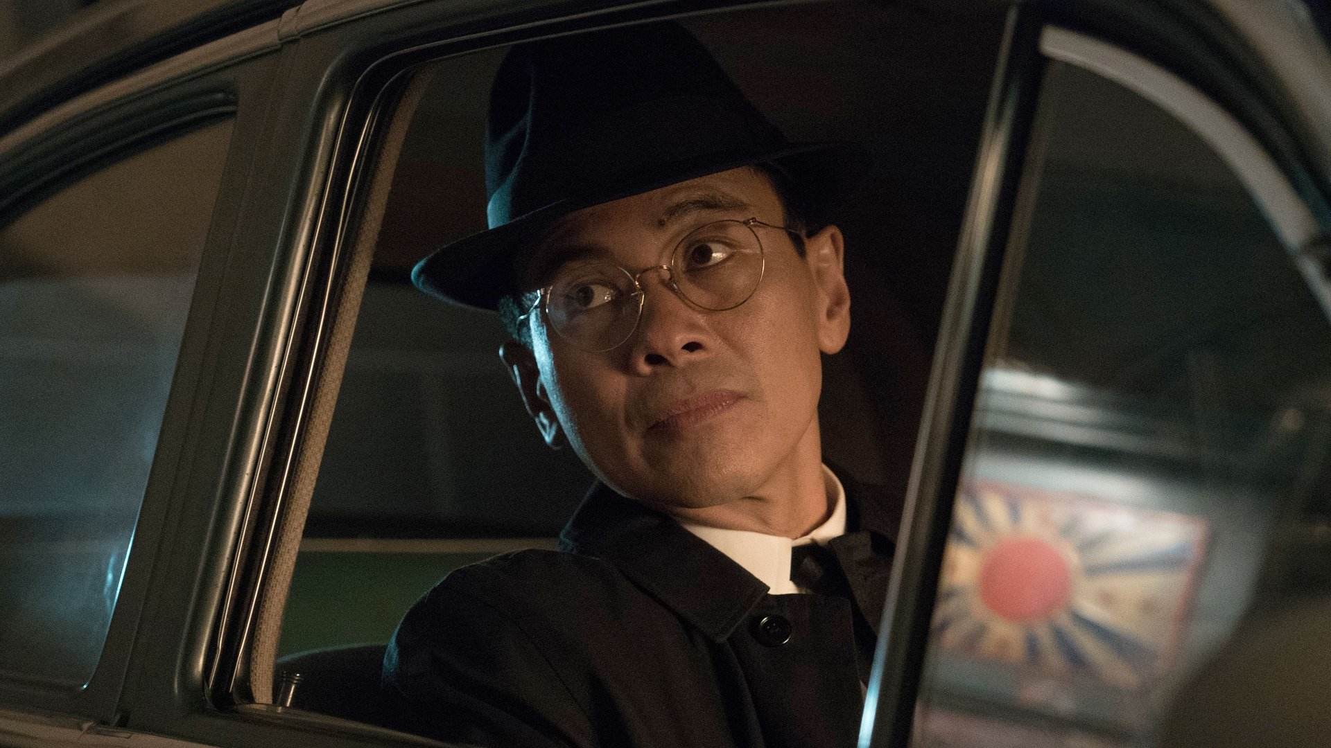 Człowiek z Wysokiego Zamku The Man in the High Castle kadr | film.org.pl Kadr z serialu Człowiek z Wysokiego Zamku: mężczyzna w kapeluszu i okularach patrzy przez okno samochodu, za nim kolorowa tarcza z czerwonym kołem. The Future is Ours