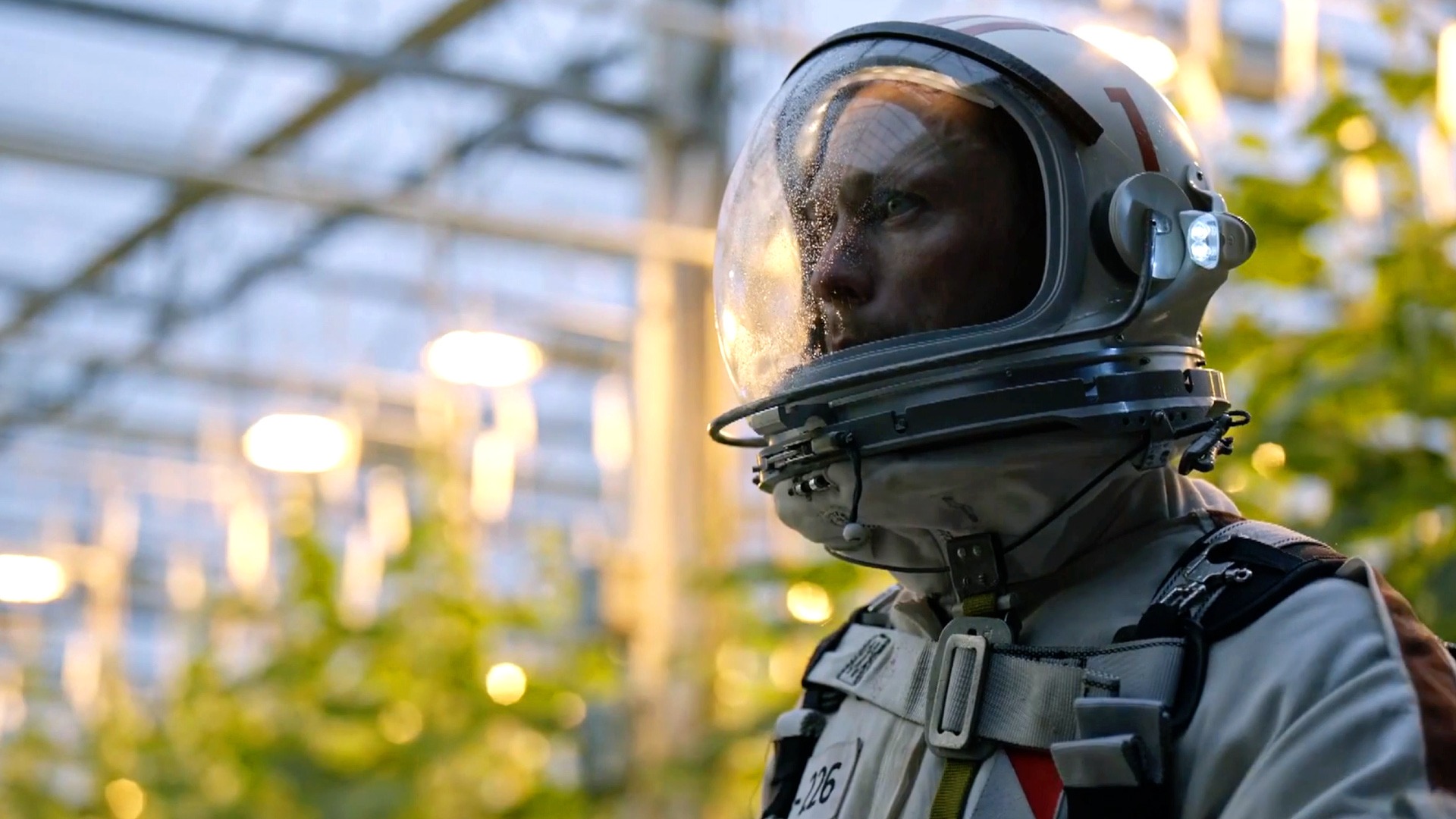 Obcy element | film.org.pl Astronaut in full suit and helmet, profile view, indoors with blurred lights and greenery in the background. Obcy element