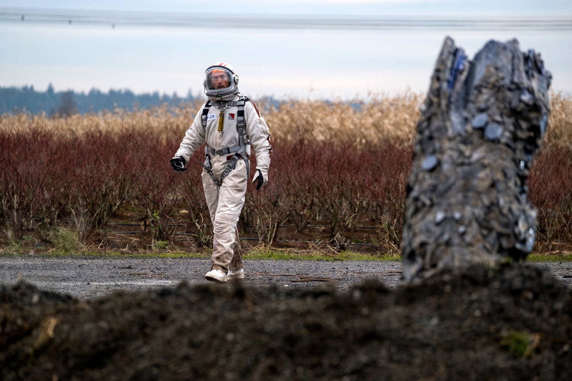 debris OBCY ELEMENT | film.org.pl Astronaut in a white spacesuit walking through a field of tall brush, with a rocky formation on the right. debris OBCY ELEMENT
