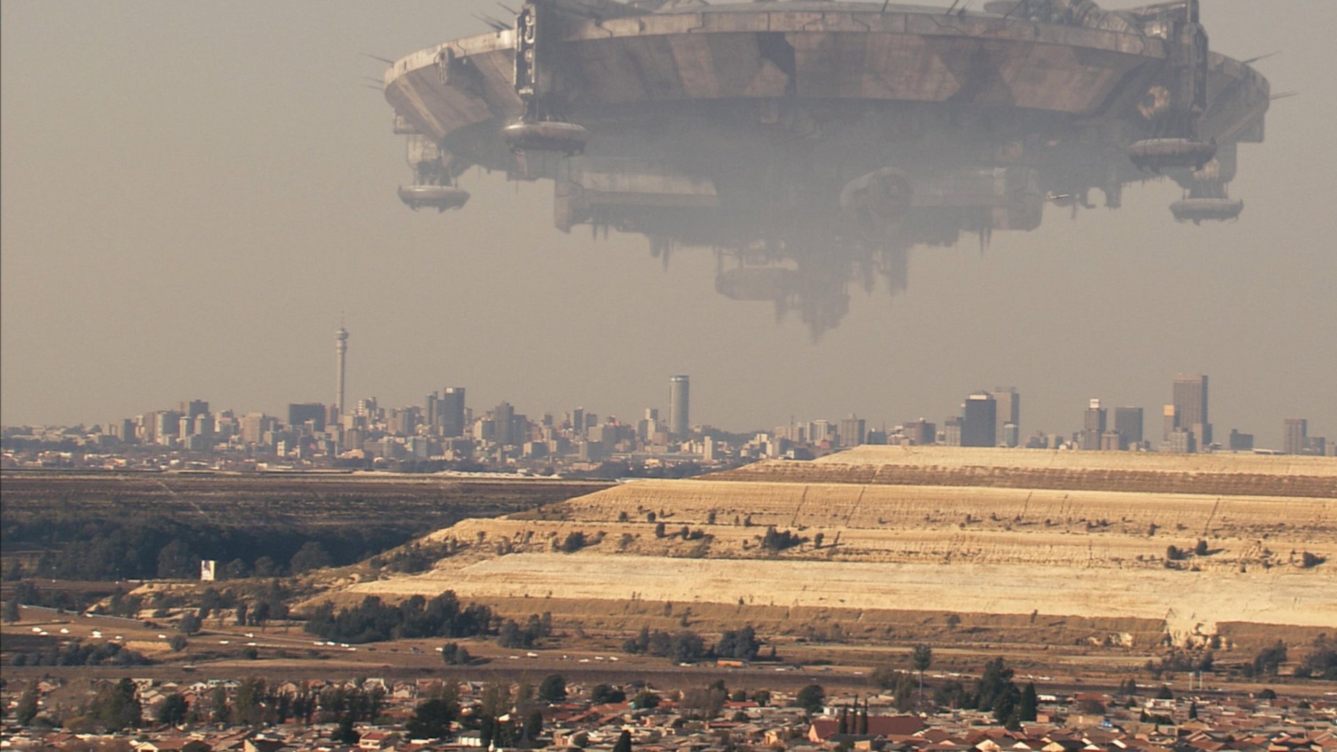 Dystrykt 9 District 9 kadr | film.org.pl Kadr z filmu Dystrykt 9: ogromny statek kosmiczny unosi się nad miastem, pod nim suchy teren i zabudowania mieszkalne.