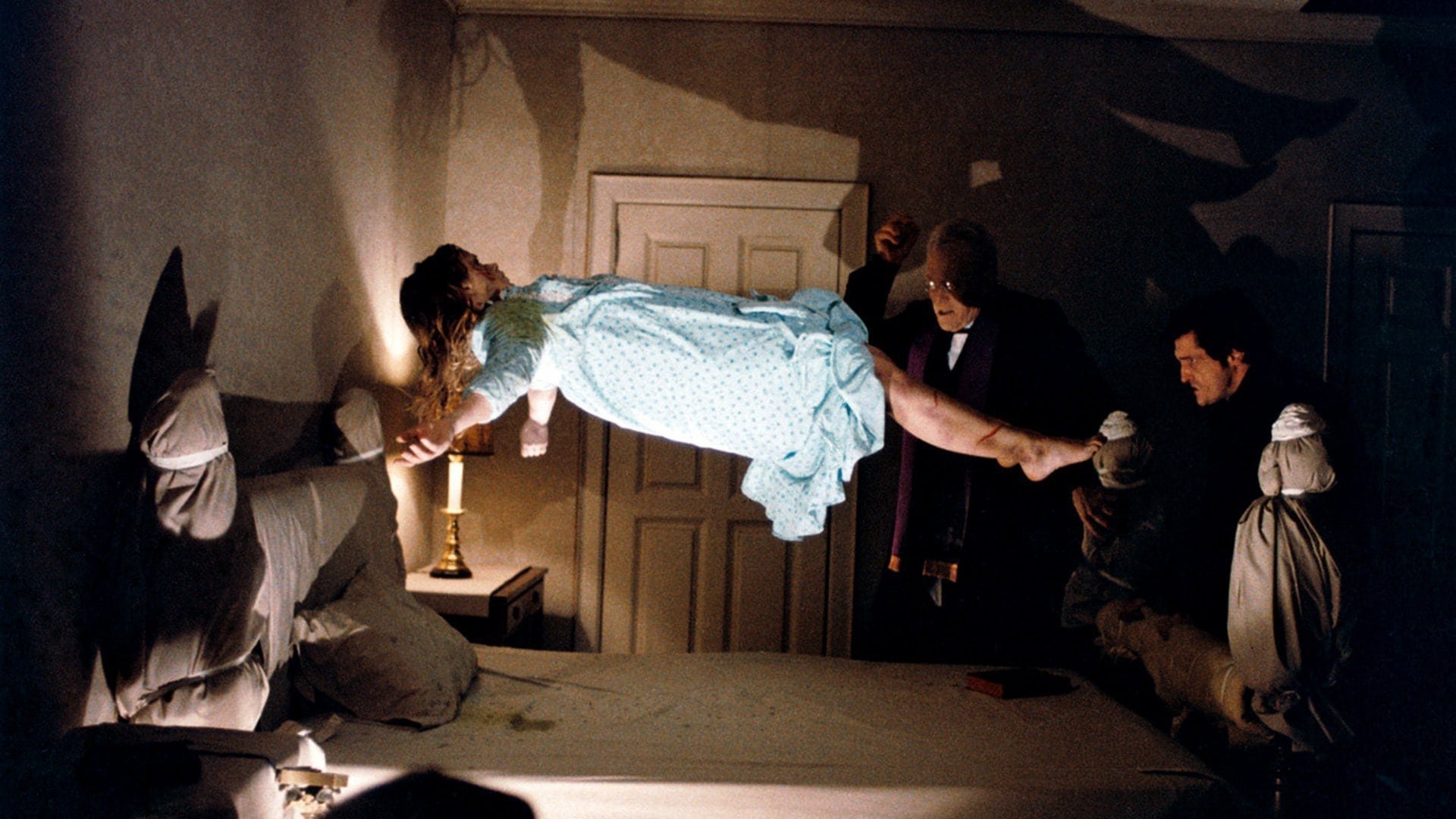 Egzorcysta The Exorcist kadr | film.org.pl Kadr z filmu Egzorcysta: dziewczynka w niebieskiej nocnej koszuli unosi się w powietrzu nad łóżkiem, obok zapalona świeca i mężczyźni w ciemnych ubraniach.