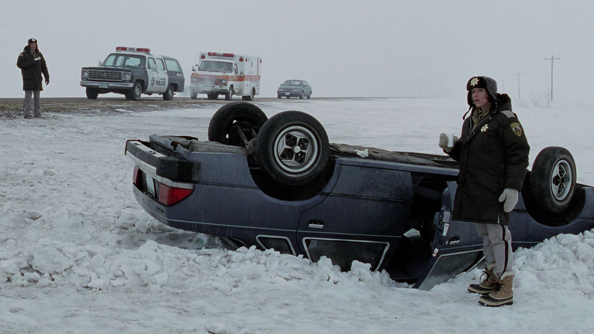 Kadr z filmu Fargo: przewrócony granatowy samochód w zaśnieżonym polu, obok umundurowany funkcjonariusz, w tle radiowóz i karetka.