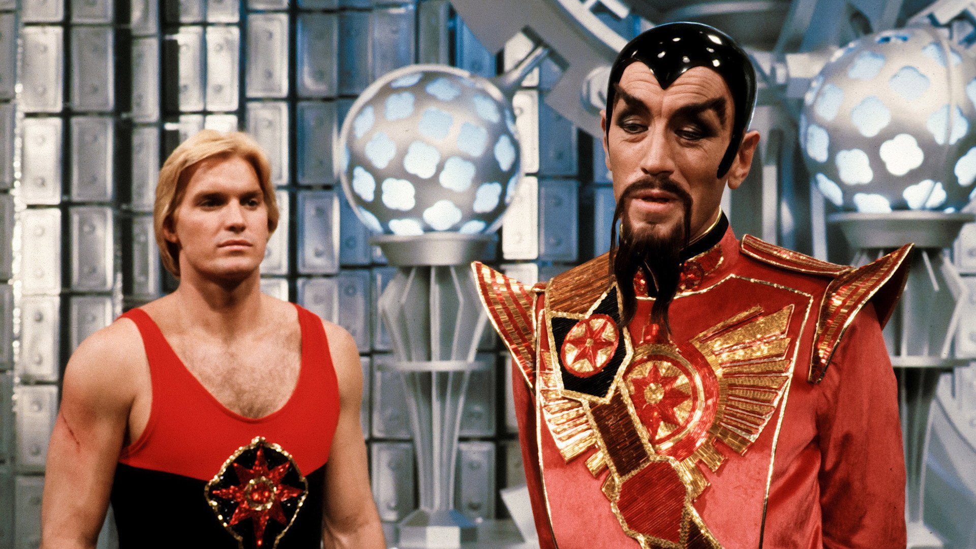 Flash Gordon kadr | film.org.pl Kadr z filmu Flash Gordon: blondwłosy mężczyzna w czerwono-czarnym stroju obok mężczyzny w czerwonym kostiumie ze złotymi zdobieniami, w metalowym wnętrzu z kulistymi lampami. space opera