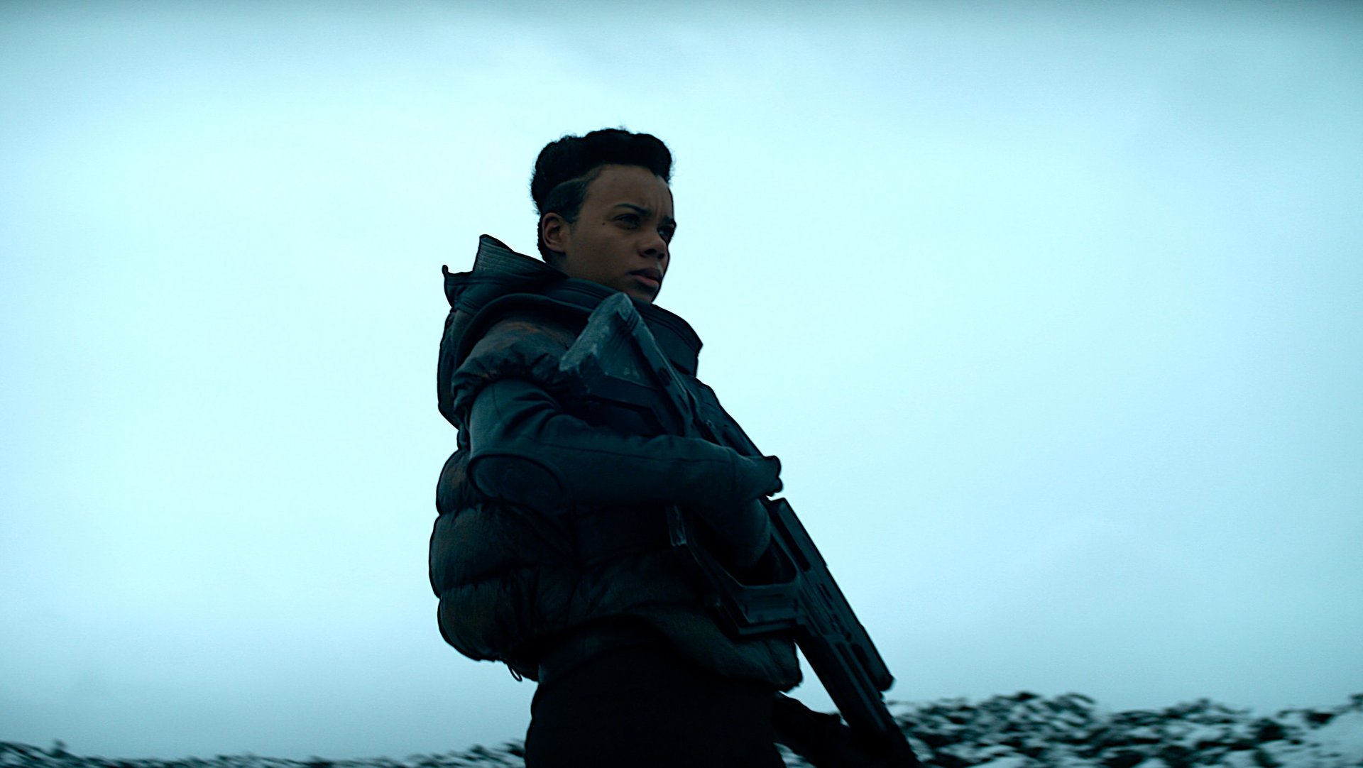 Fundacja Foundation kadr | film.org.pl Kadr z serialu Fundacja: postać w ciemnej kurtce ze stojącym kołnierzem trzyma karabin na tle pochmurnego, błękitnego nieba.