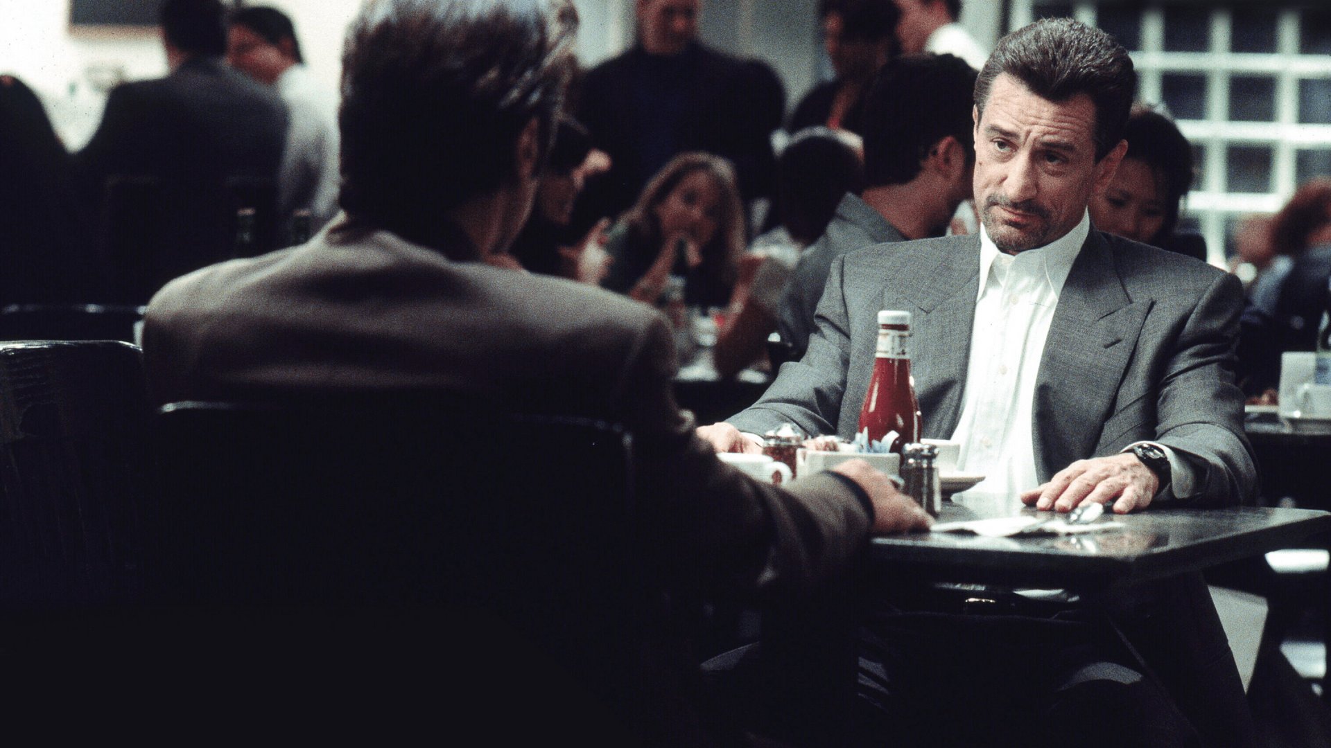 Kadr z filmu Gorączka: mężczyzna w szarym garniturze siedzi przy stoliku w kawiarni, naprzeciwko mężczyzny tyłem do kamery filmy kryminalne