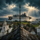 Stormy coastal town scene with a rusted iron cross on the church roof in the foreground and a rocky island lighthouse in the distance under dramatic clouds.