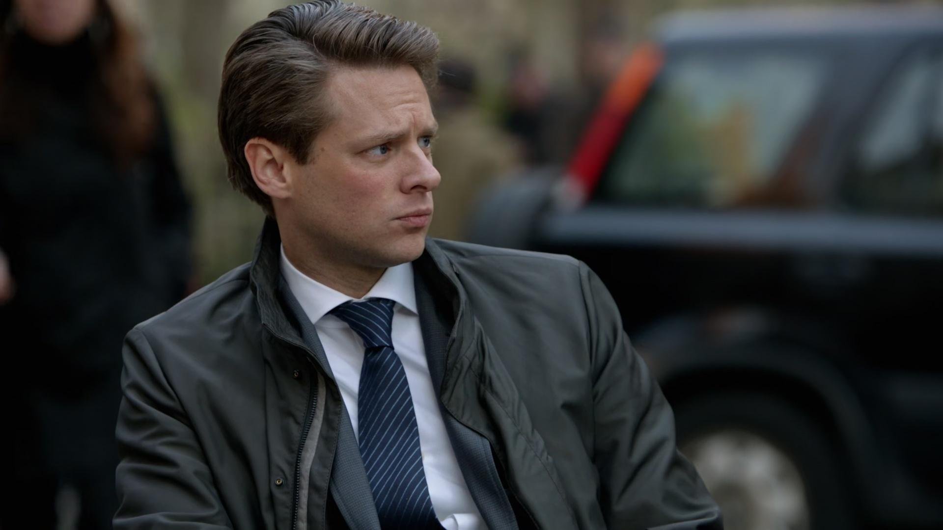 Impersonalni Person of Interest kadr | film.org.pl mężczyzna w ciemnej kurtce i granatowym krawacie w paski patrzy w bok na zewnątrz, w tle czarny samochód.