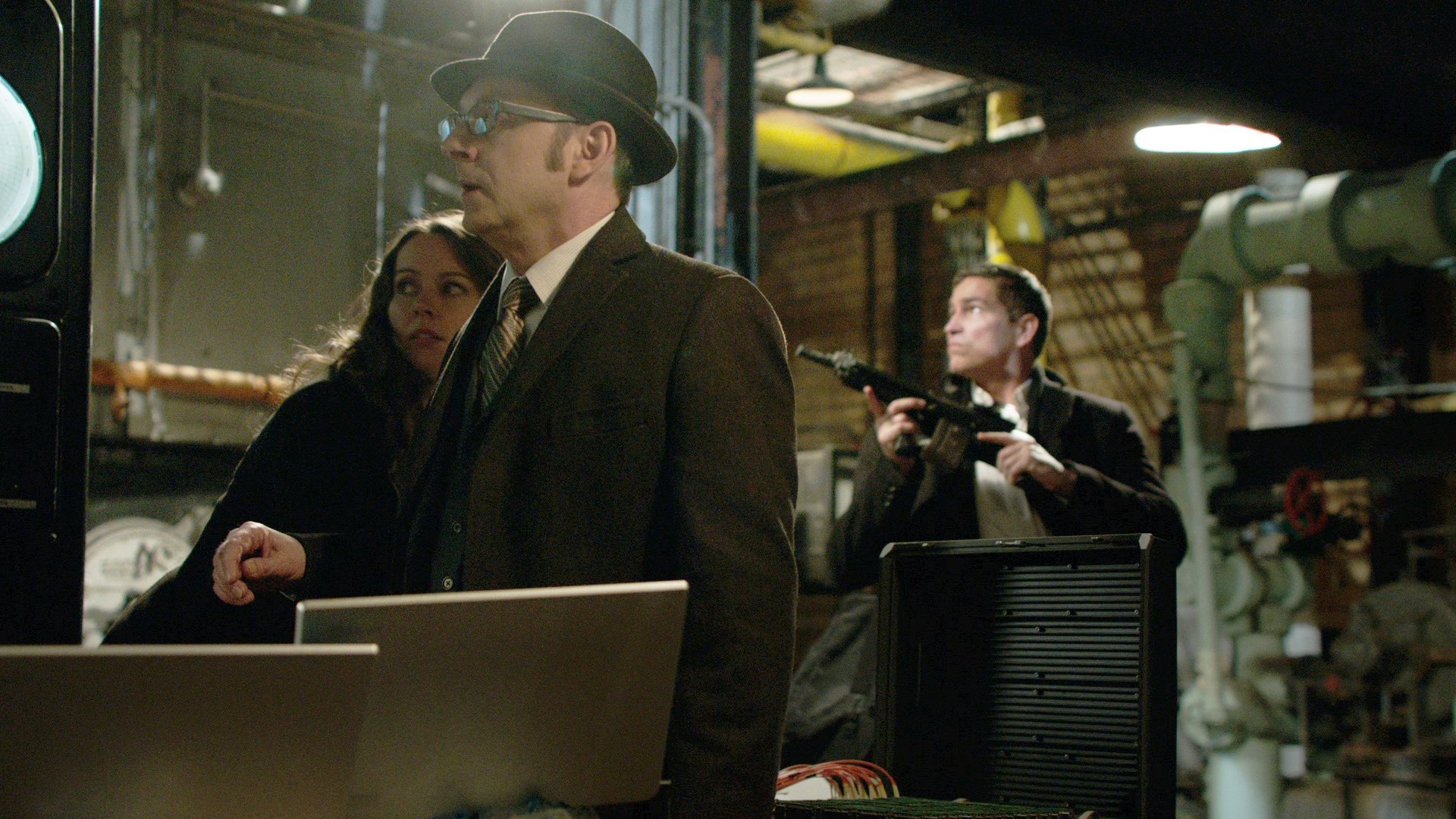 Impersonalni Person of Interest kadr | film.org.pl Kadr z serialu Impersonalni: mężczyzna w kapeluszu i okularach stoi przy laptopie, obok kobieta, w tle uzbrojony mężczyzna w industrialnym wnętrzu.