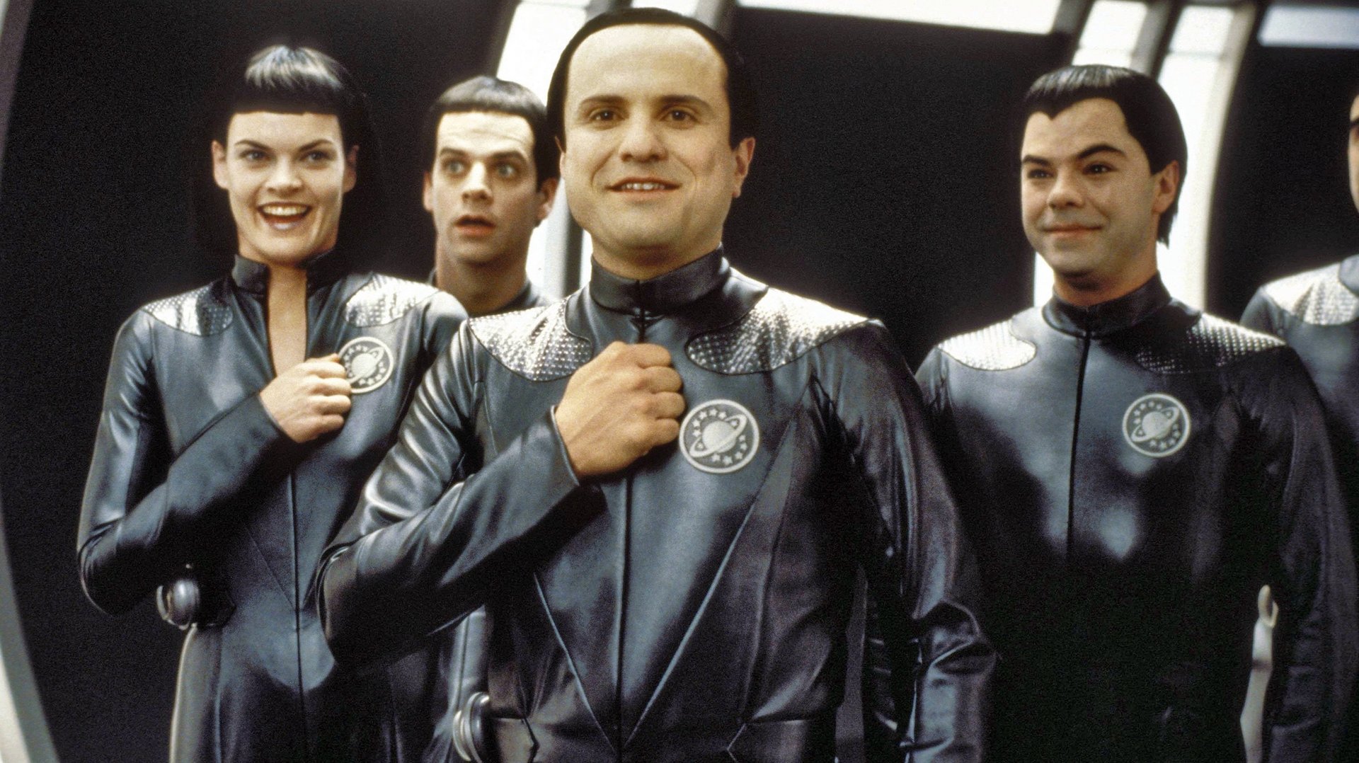 Kosmiczna załoga Galaxy Quest kadr | film.org.pl Kadr z serialu Kosmiczna załoga: czworo ludzi w czarnych futurystycznych kombinezonach z emblematem na piersi, stoi w metalicznym wnętrzu statku.