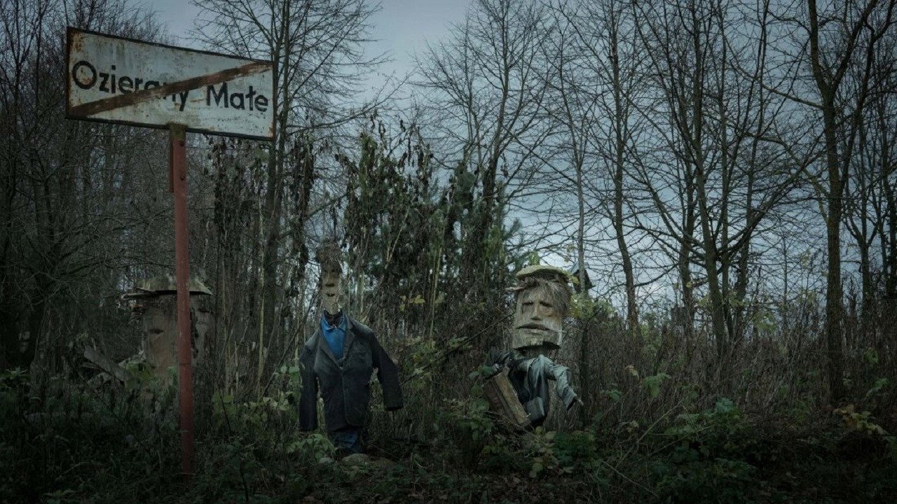 Kruk kadr | film.org.pl Kadr z serialu Kruk. Szepty słychać po zmroku: tablica z napisem Ozierany Małe i dwie rzeźbione figury w zaroślach.