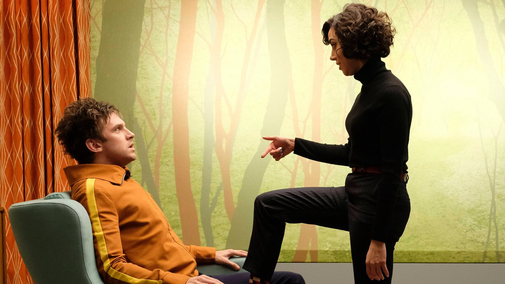 Legion kadr | film.org.pl Kadr z serialu Legion: mężczyzna w pomarańczowej bluzie siedzi w fotelu, kobieta w czarnym golfie wskazuje na niego palcem.