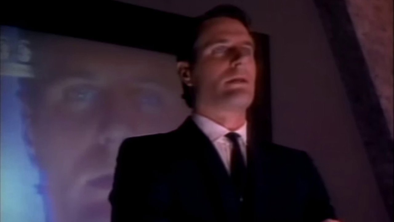 Kadr z serialu Max Headroom: mężczyzna w ciemnym garniturze i krawacie stoi w mroku przed jasnym ekranem z twarzą.