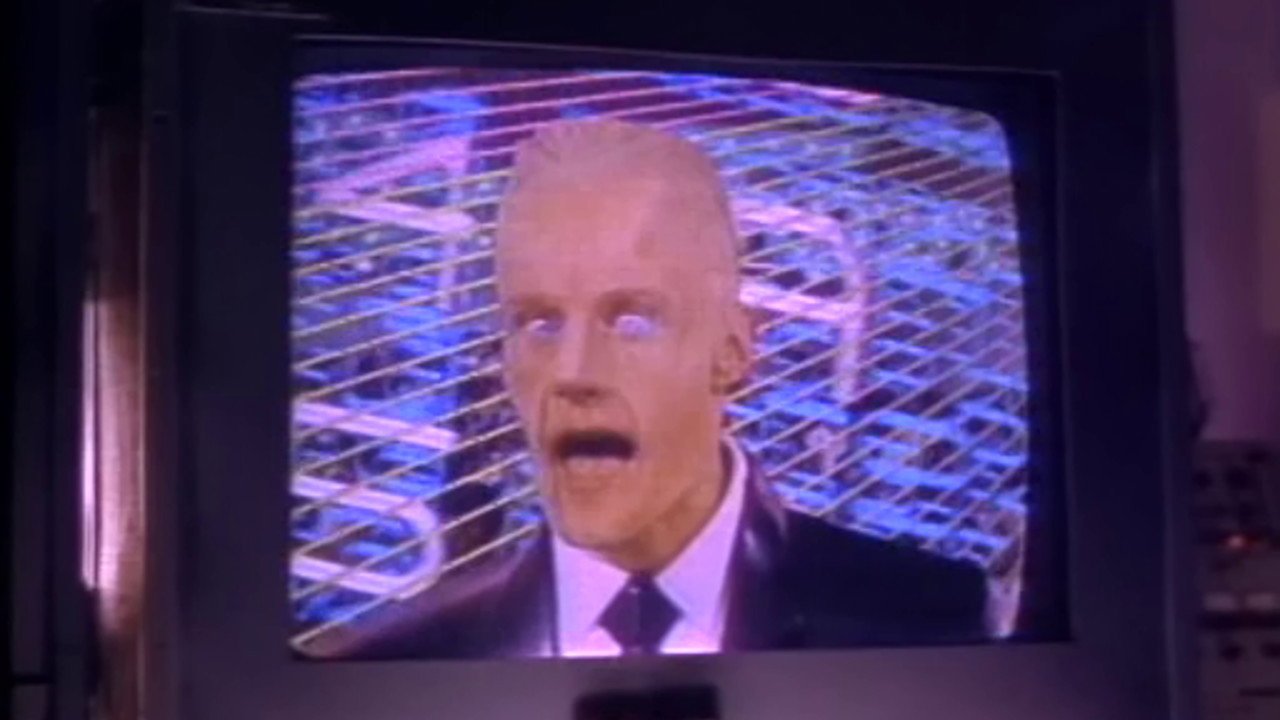 Kadr z serialu Max Headroom: na ekranie telewizora blady mężczyzna w garniturze z otwartymi ustami, za nim niebieska siatka.