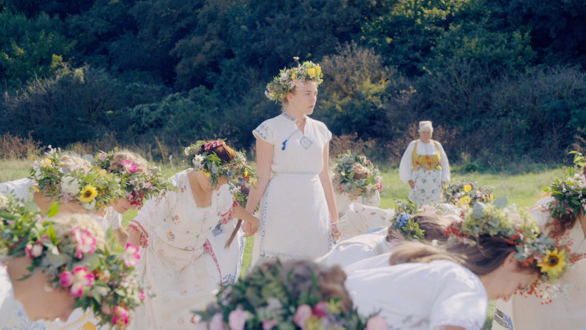 Midsommar W biały dzień Midsommar kadr | film.org.pl Kadr z filmu Midsommar. W biały dzień: w słonecznym krajobrazie stoi kobieta w białej sukni z wiankiem z kwiatów, a wokół kłaniają się uczestnicy w kwiecistych wiankach.