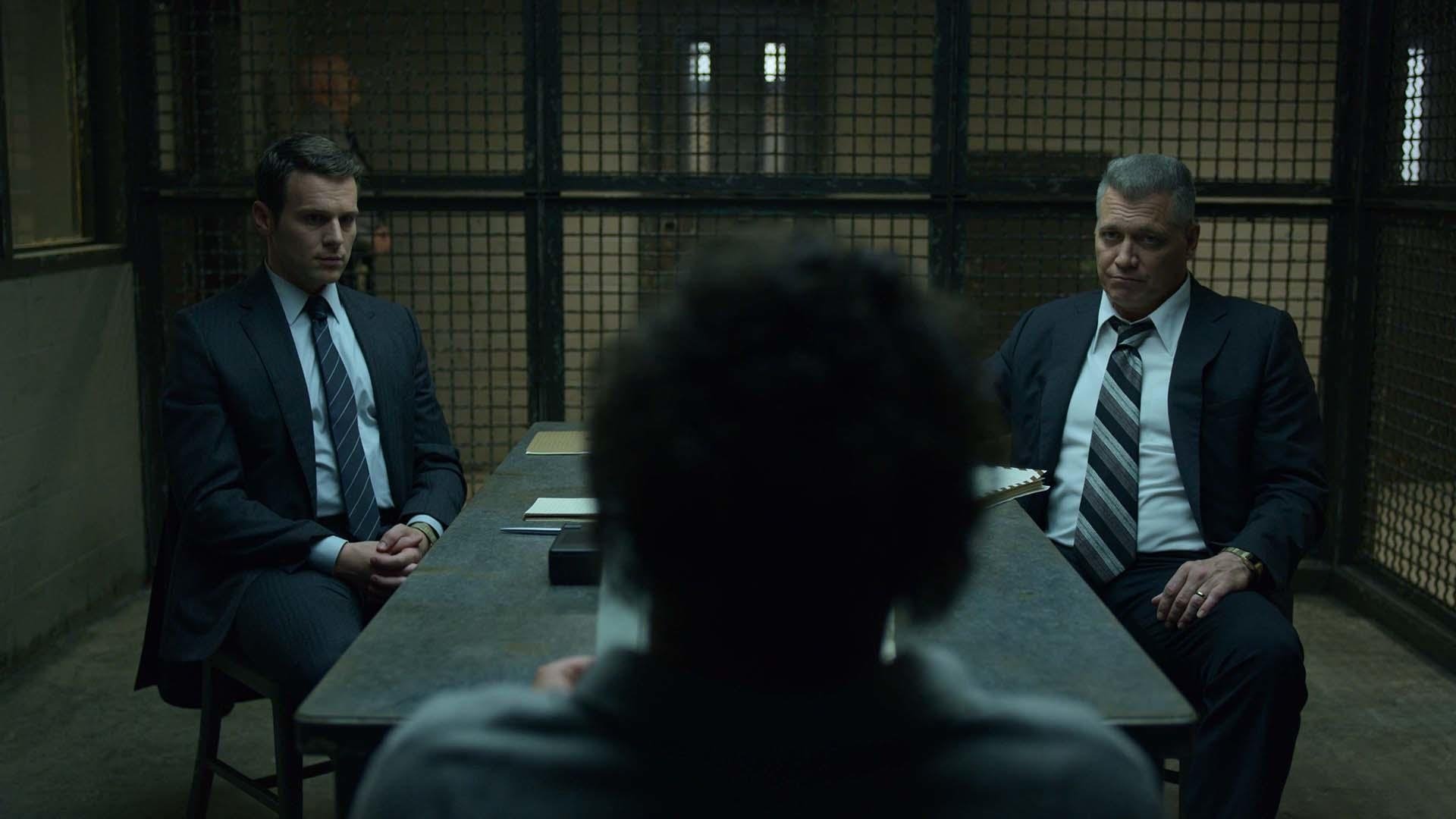 MINDHUNTER kadr | film.org.pl Kadr z serialu Mindhunter: dwóch mężczyzn w garniturach siedzi po obu stronach stołu w zakratowanym pomieszczeniu, naprzeciw nich tyłem stoi osoba w ciemnej bluzie. True Detective