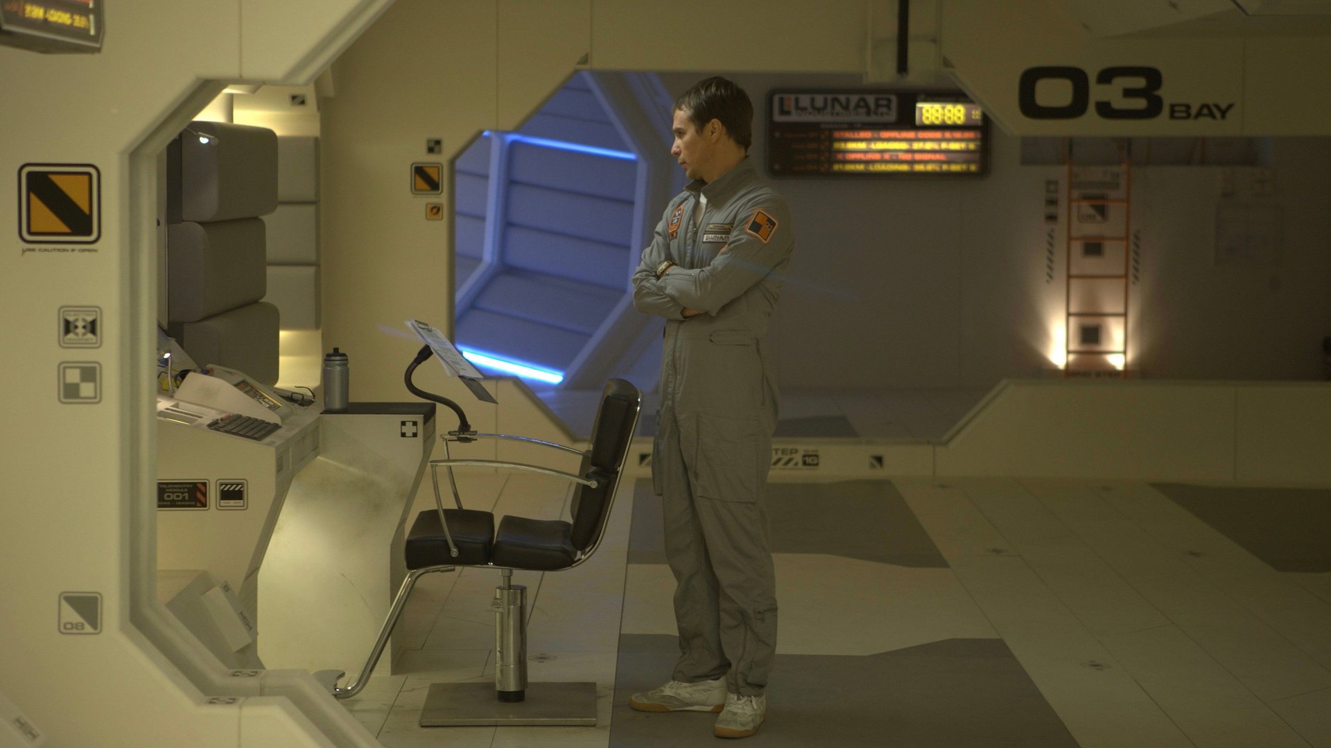 Moon kadr | film.org.pl Kadr z filmu Moon: mężczyzna w szarym kombinezonie stoi z założonymi rękami w futurystycznym wnętrzu z fotelem i konsolą Thriller sci-fi