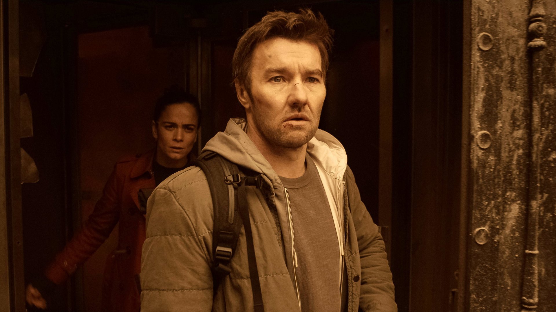 Mroczna materia Dark Matter kadr | film.org.pl Kadr z serialu Mroczna materia: mężczyzna w szarej kurtce z plecakiem patrzy przed siebie, za nim stoi kobieta w czerwonej kurtce.