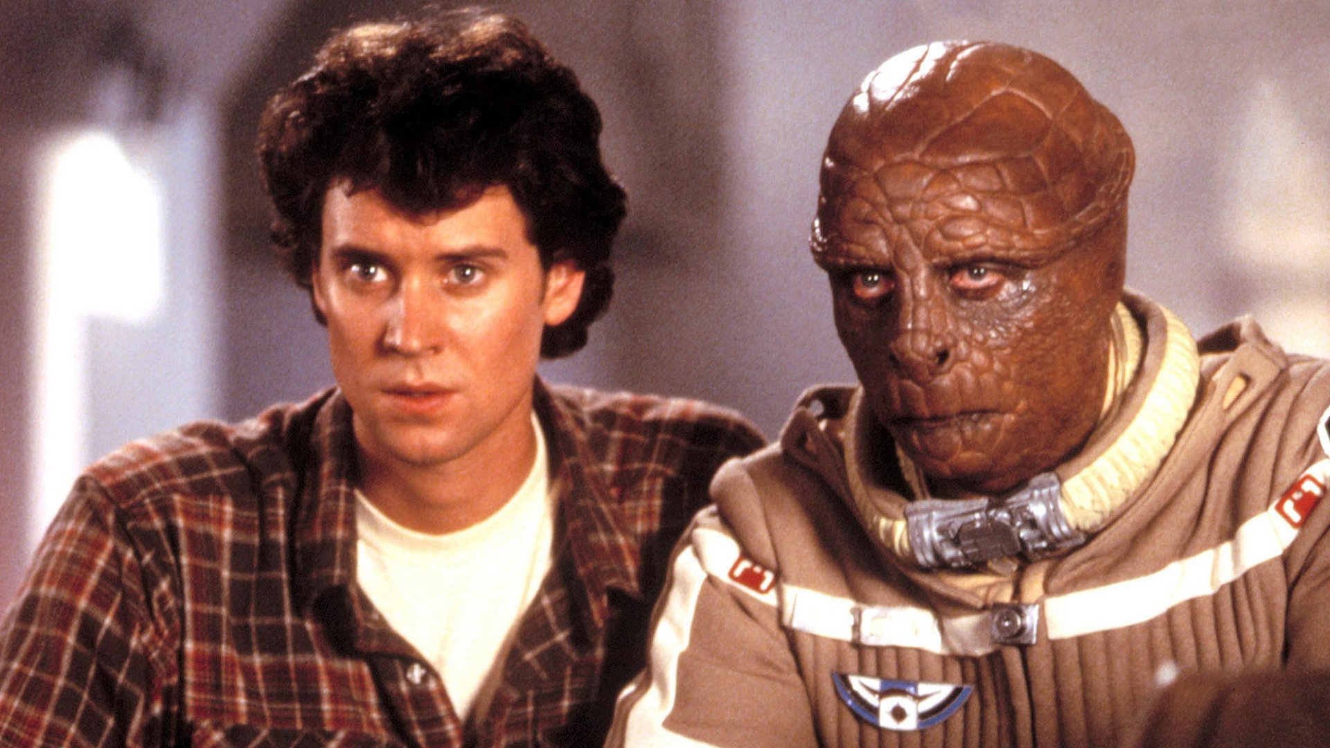 Ostatni gwiezdny wojownik The Last Starfighter kadr | film.org.pl Kadr z filmu Ostatni gwiezdny wojownik: młody mężczyzna w kraciastej koszuli obok istoty z gadzią, brązową twarzą w beżowym kombinezonie. space opera