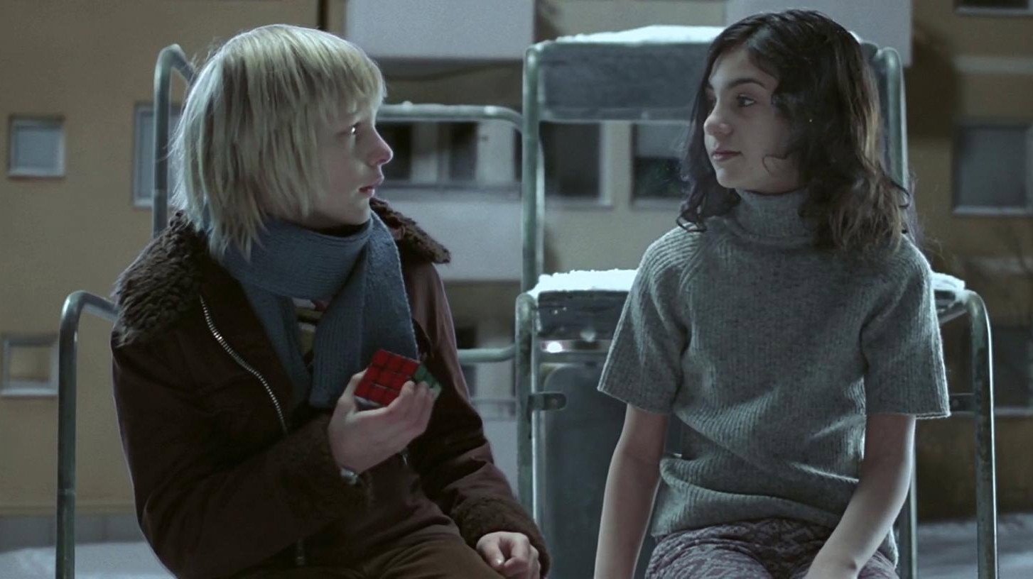 Pozwól mi wejść Låt den rätte komma in kadr | film.org.pl Kadr z filmu Pozwól mi wejść: blond chłopiec w brązowej kurtce i szaliku trzyma czerwone pudełko, obok ciemnowłosa dziewczynka w szarym swetrze siedzi na zimowym podwórku