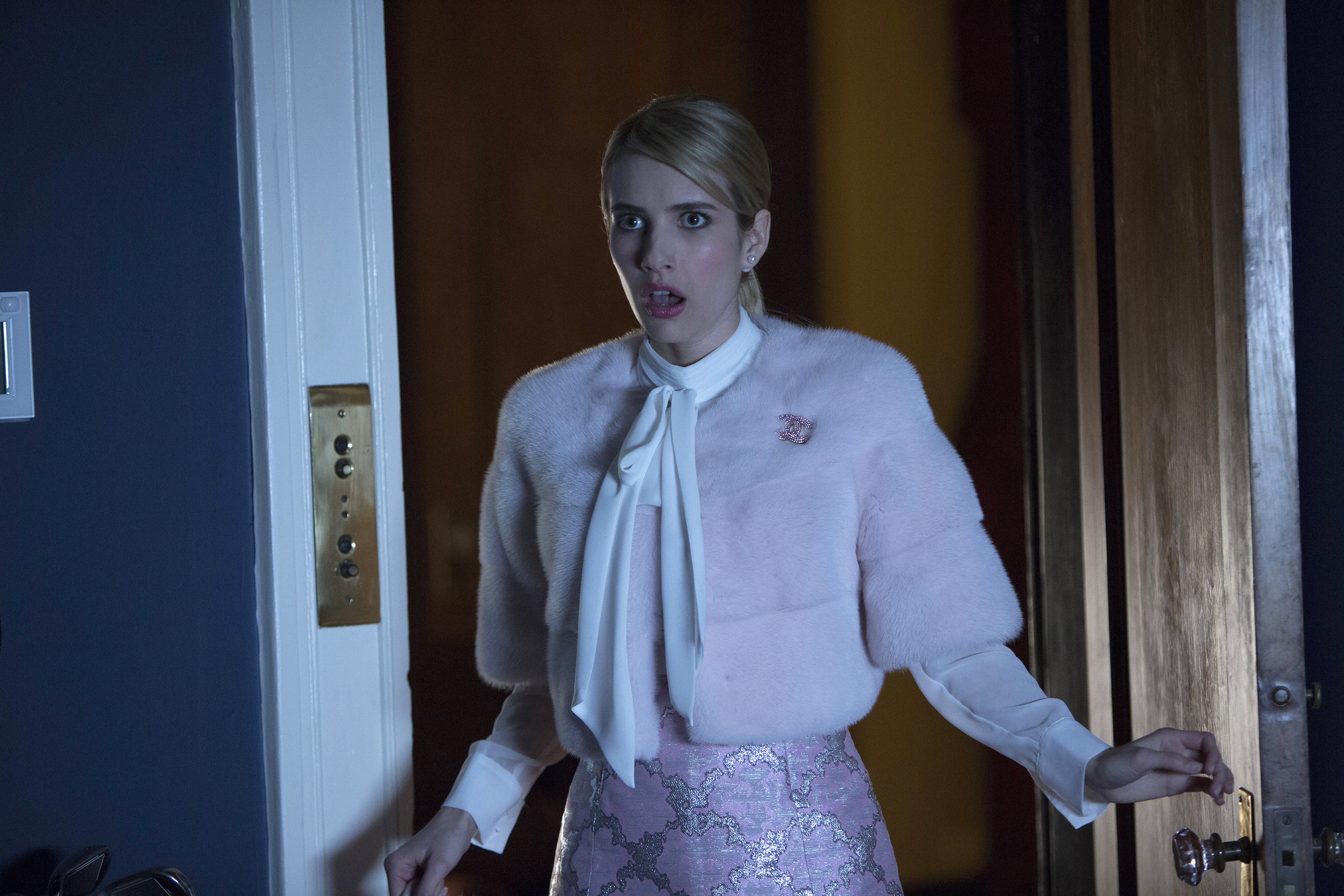 Woman with blonde hair wearing a pale pink fuzzy cardigan and white blouse, standing in a doorway with a surprised expression. screem queens krolowe krzyku