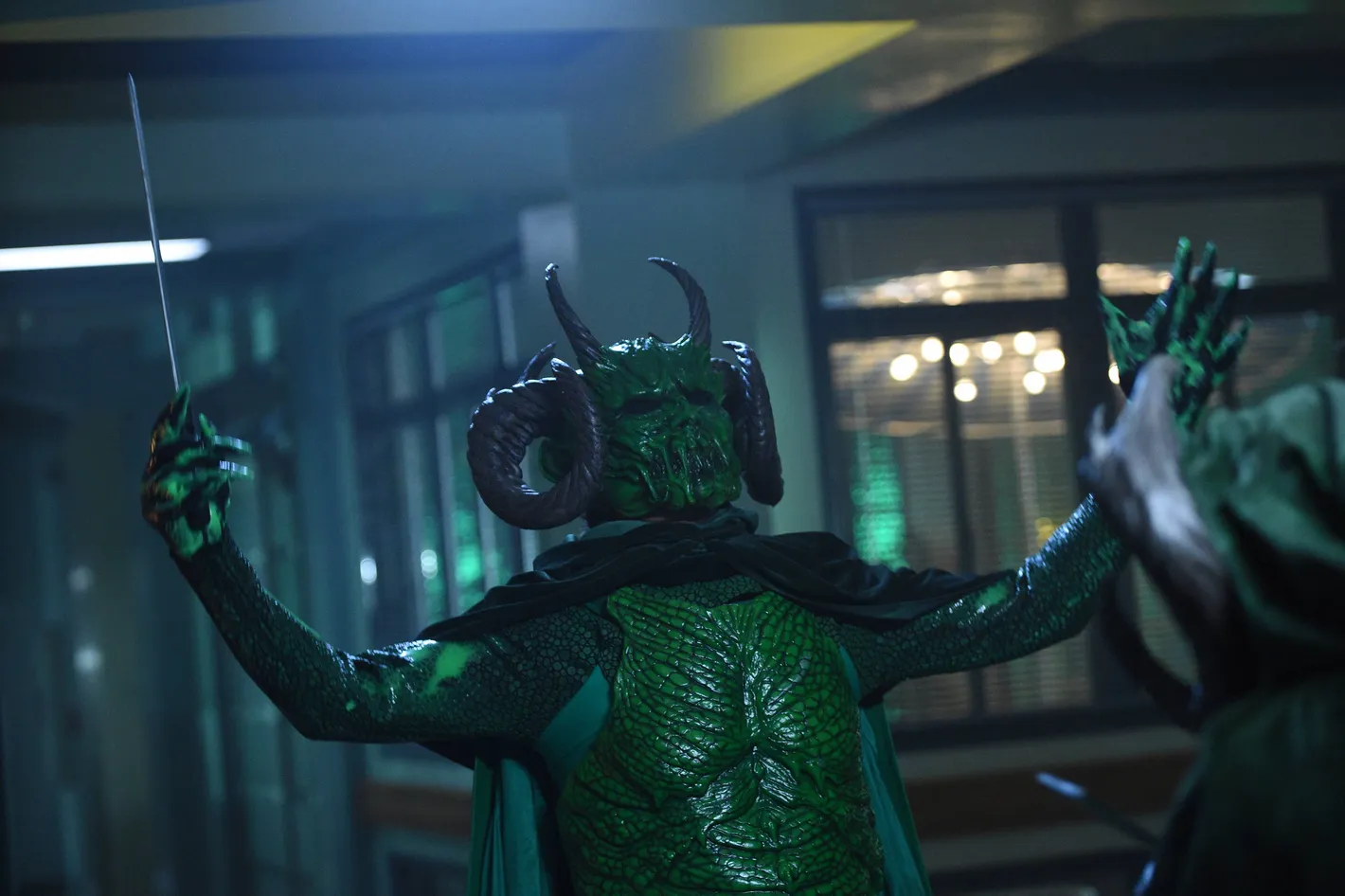 Green dragon-costumed character with large curved horns and textured scales raises a sword in a dim indoor hallway (cosplay scene).