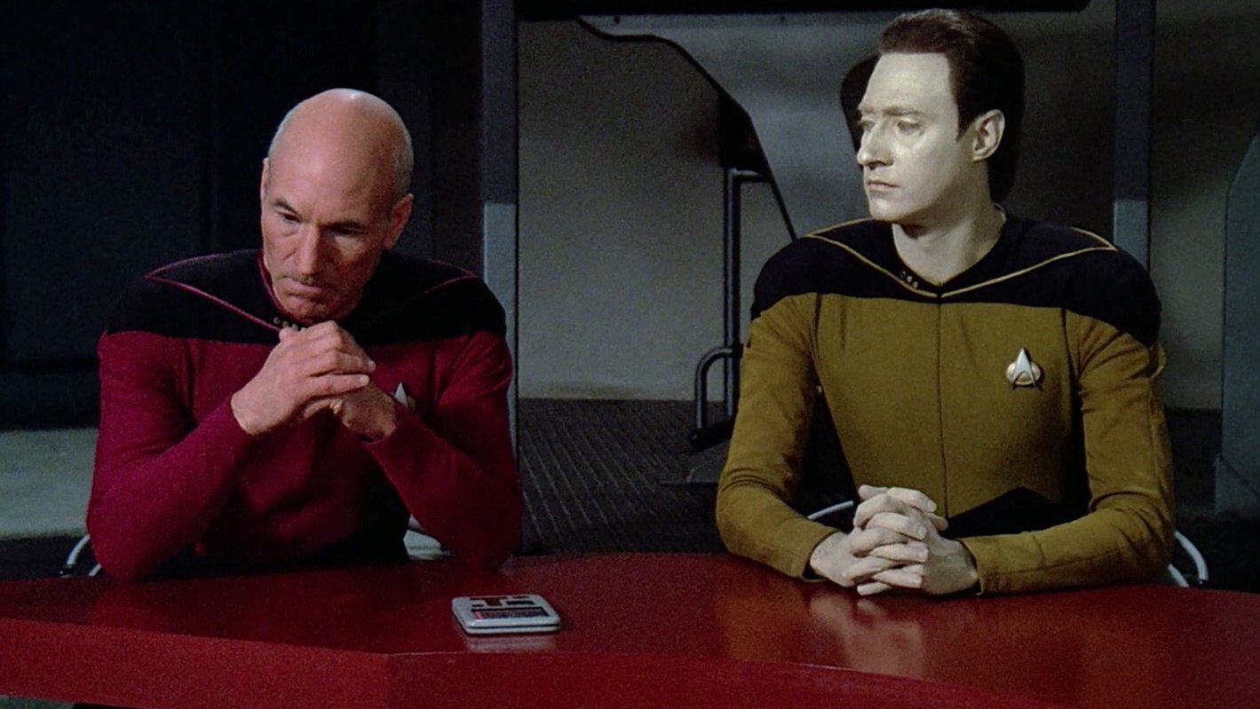 Kadr z serialu Star Trek: Następne pokolenie: dwaj mężczyźni w czerwonym i złotym mundurze siedzą przy czerwonym stole. seriale sci-fi