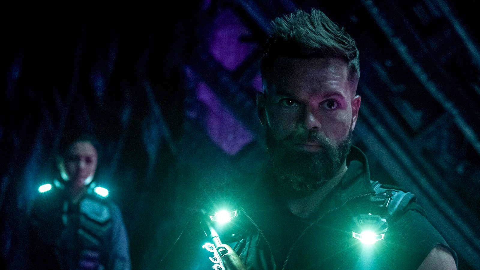 Kadr z serialu The Expanse: brodaty mężczyzna w ciemnym kombinezonie z zielonymi świecącymi elementami sprzętu, w tle kobieta, fioletowe oświetlenie.