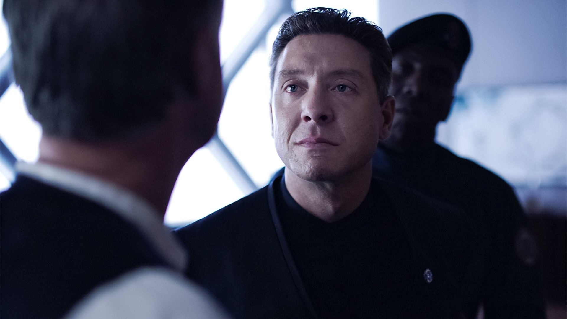 Kadr z serialu The Expanse: poważny mężczyzna w czarnym mundurze patrzy przed siebie, obok stoi drugi mężczyzna w ciemnym stroju.