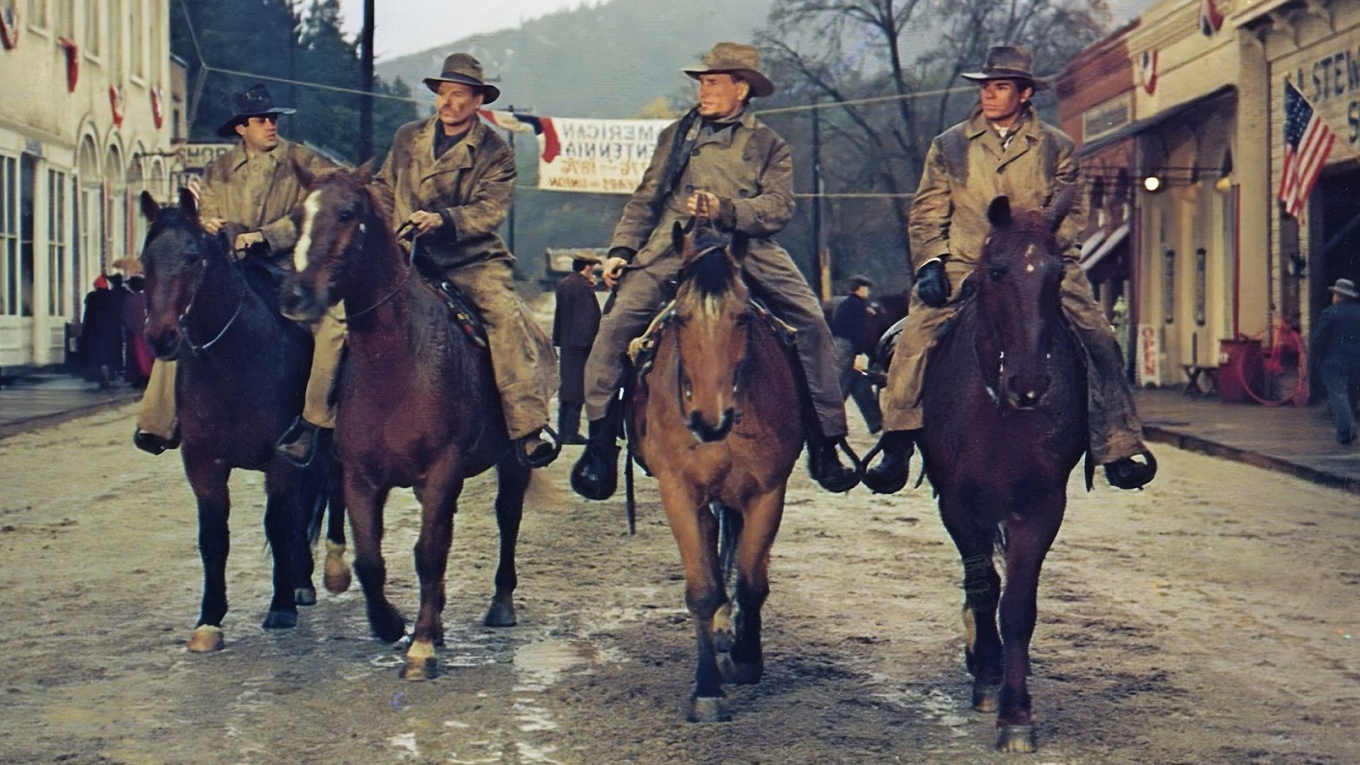 The Great Northfield Minnesota Raid kadr | film.org.pl Kadr z filmu Wielki napad w Minnesocie: czterech mężczyzn w długich płaszczach i kapeluszach jedzie konno ulicą małego miasteczka.
