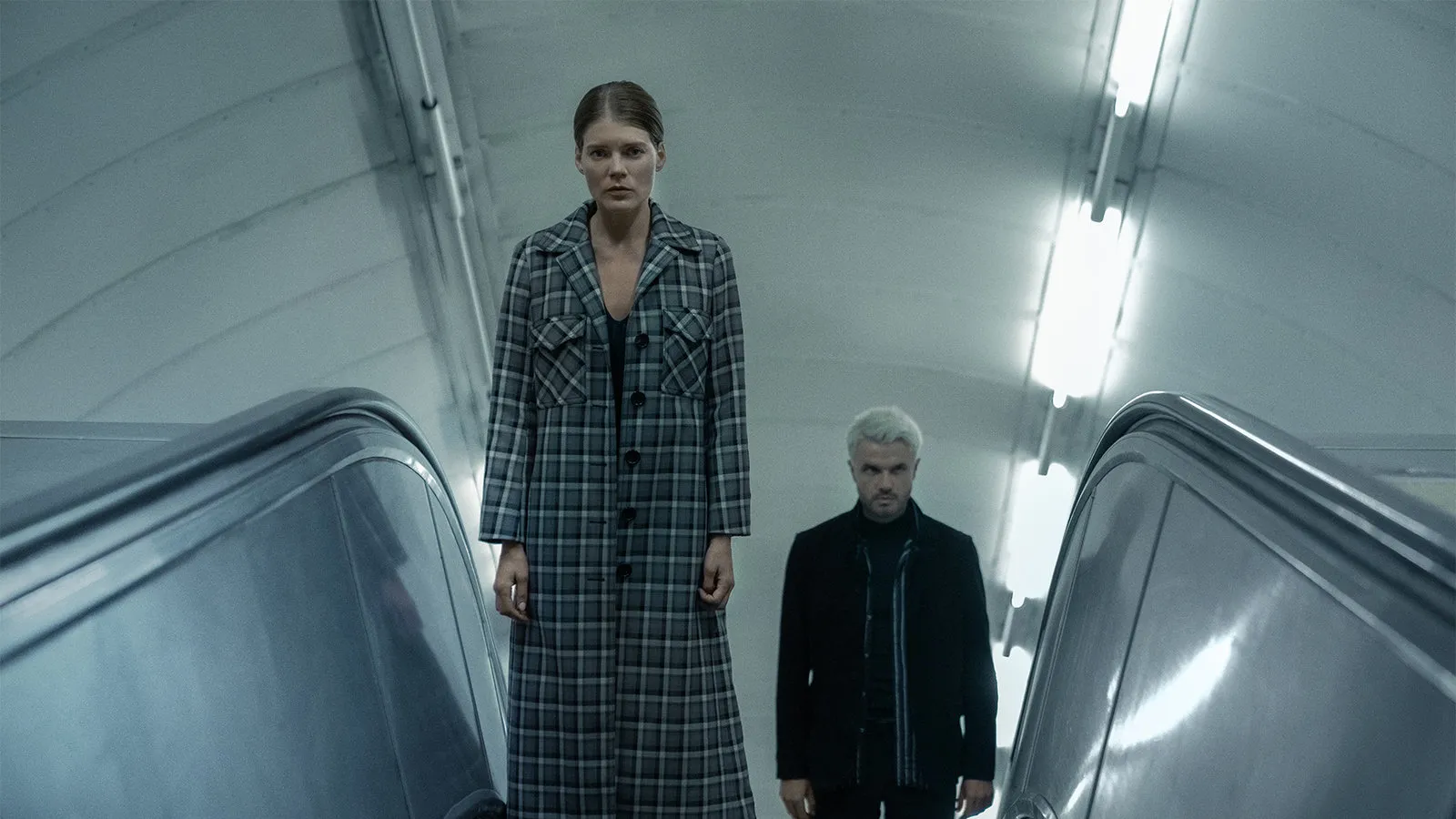 Two people on an escalator in a subway tunnel; woman in a plaid coat leads, man in a dark jacket follows behind. The Rook