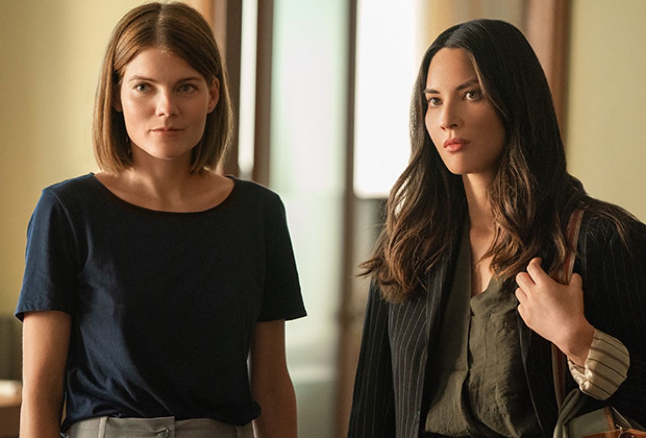 Two women stand in a sunlit hallway, one with a navy top and the other in a pinstriped blazer carrying a bag. The Rook