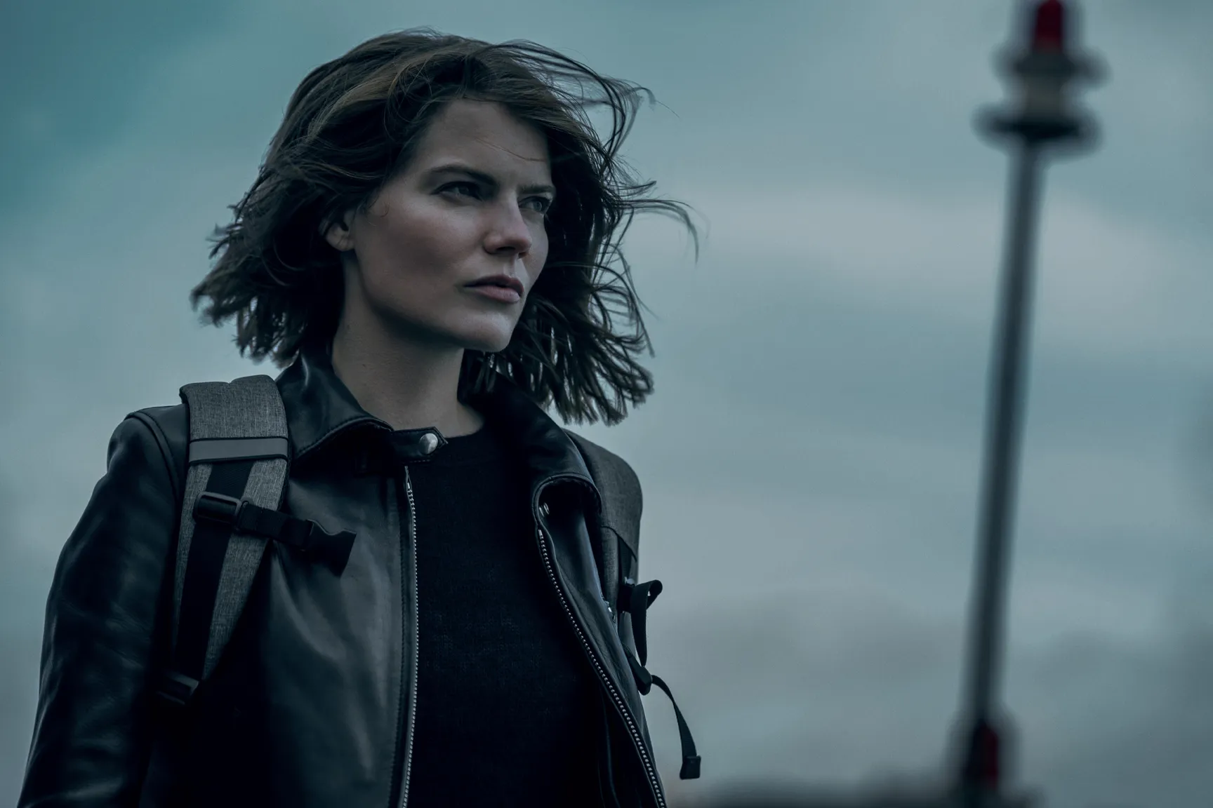 Windy coastal portrait of a woman with wind-blown hair, wearing a black leather jacket and backpack, with a distant lighthouse in the background.