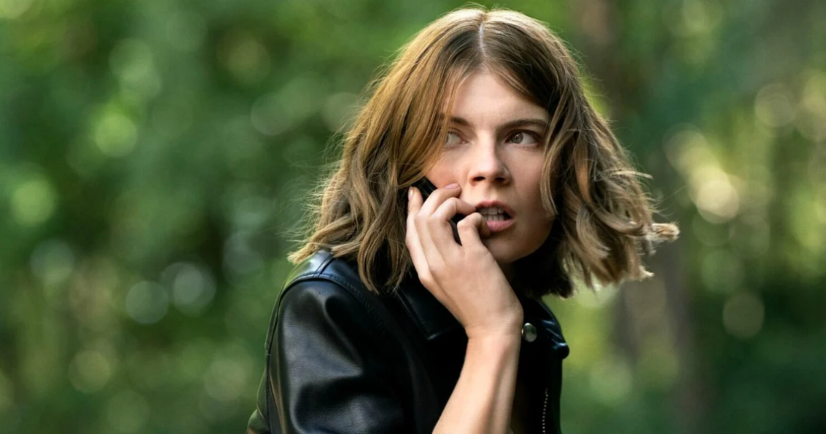 Woman in a black leather jacket holds a smartphone to her ear, looking to the side in a leafy outdoor park. The Rook