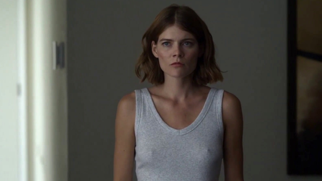 Woman with shoulder-length brown hair in a gray tank top faces the camera with a serious expression in a dim hallway. The Rook