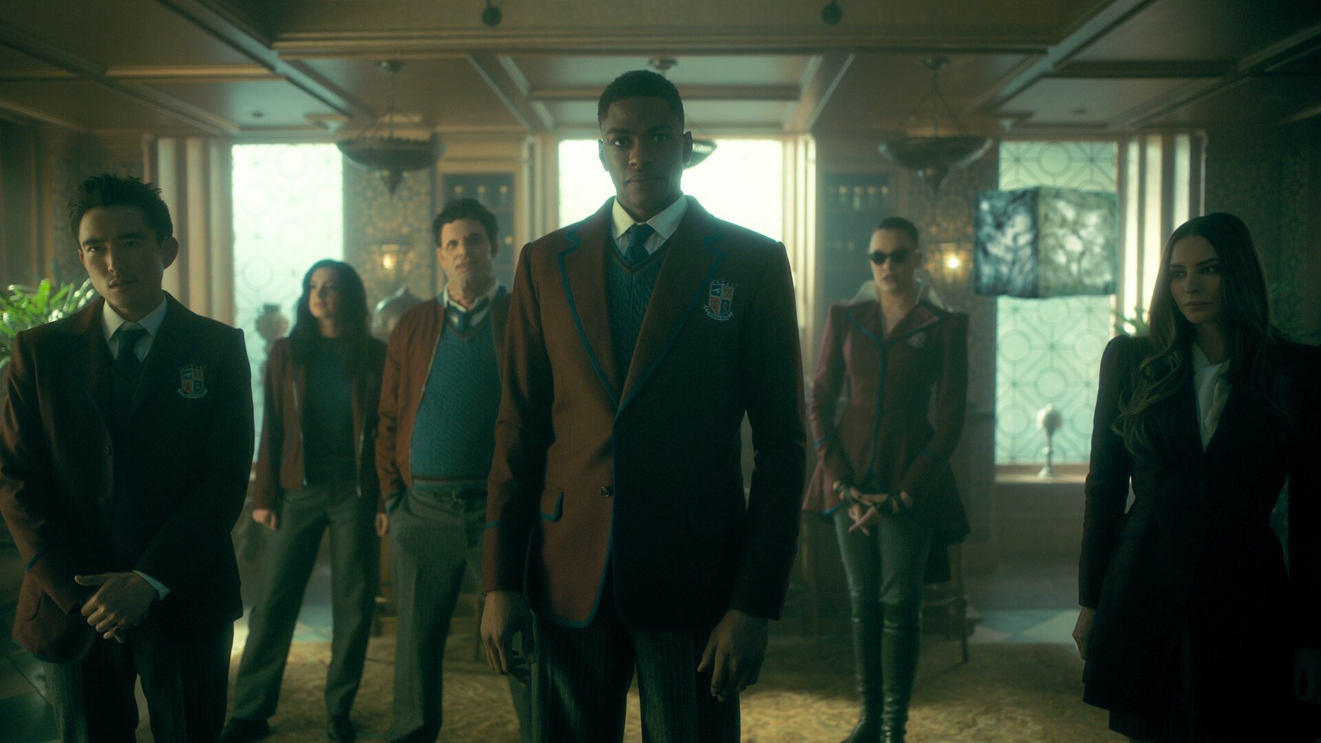 The Umbrella Academy kadr | film.org.pl Kadr z serialu The Umbrella Academy: sześcioro młodych ludzi w szkolnych marynarkach stoi w rzędzie w eleganckim wnętrzu z lampami.