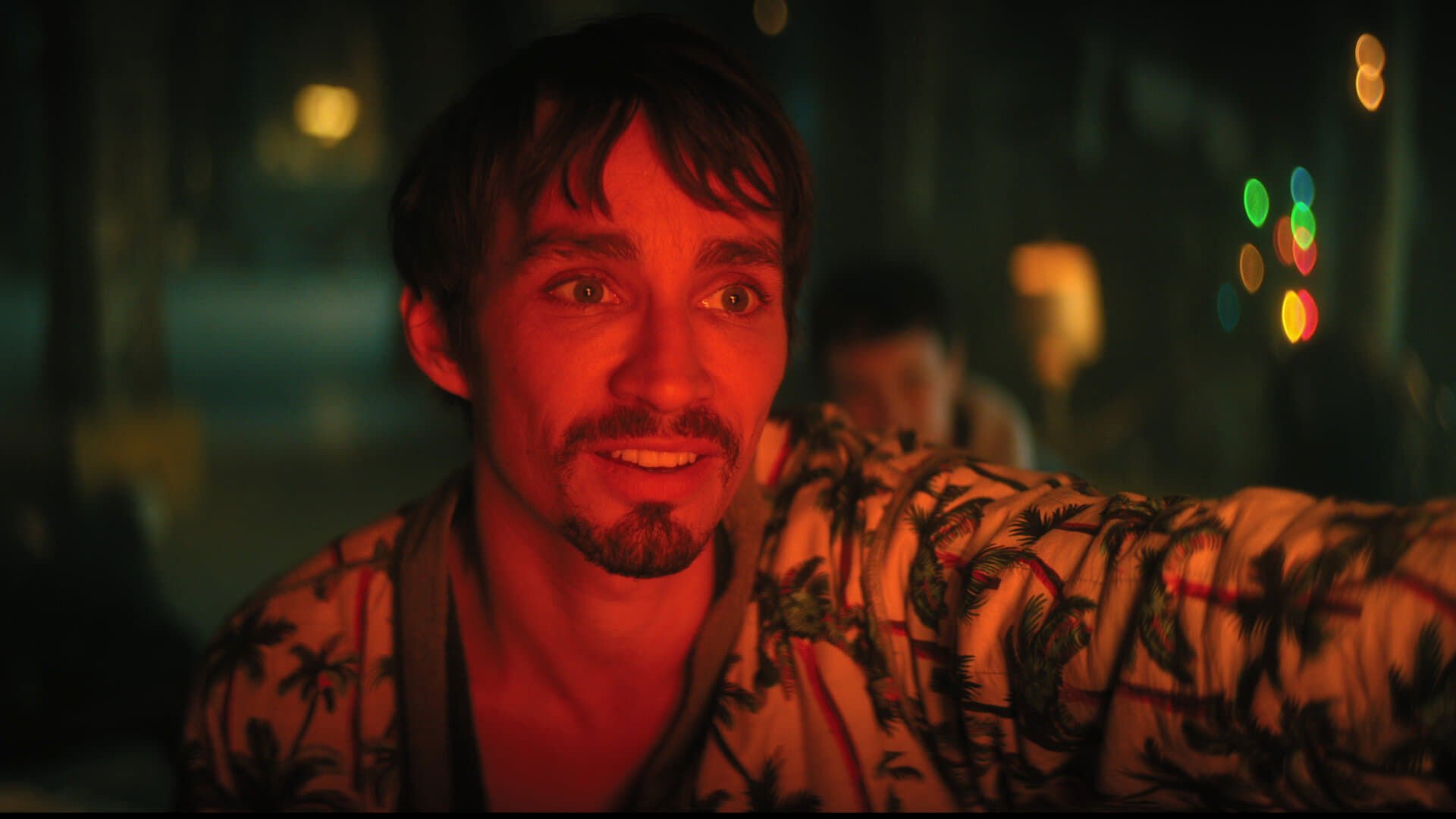 The Umbrella Academy kadr | film.org.pl Kadr z serialu The Umbrella Academy: mężczyzna z brodą w koszuli z palmowym wzorem, czerwone oświetlenie, rozmazane światła w tle.