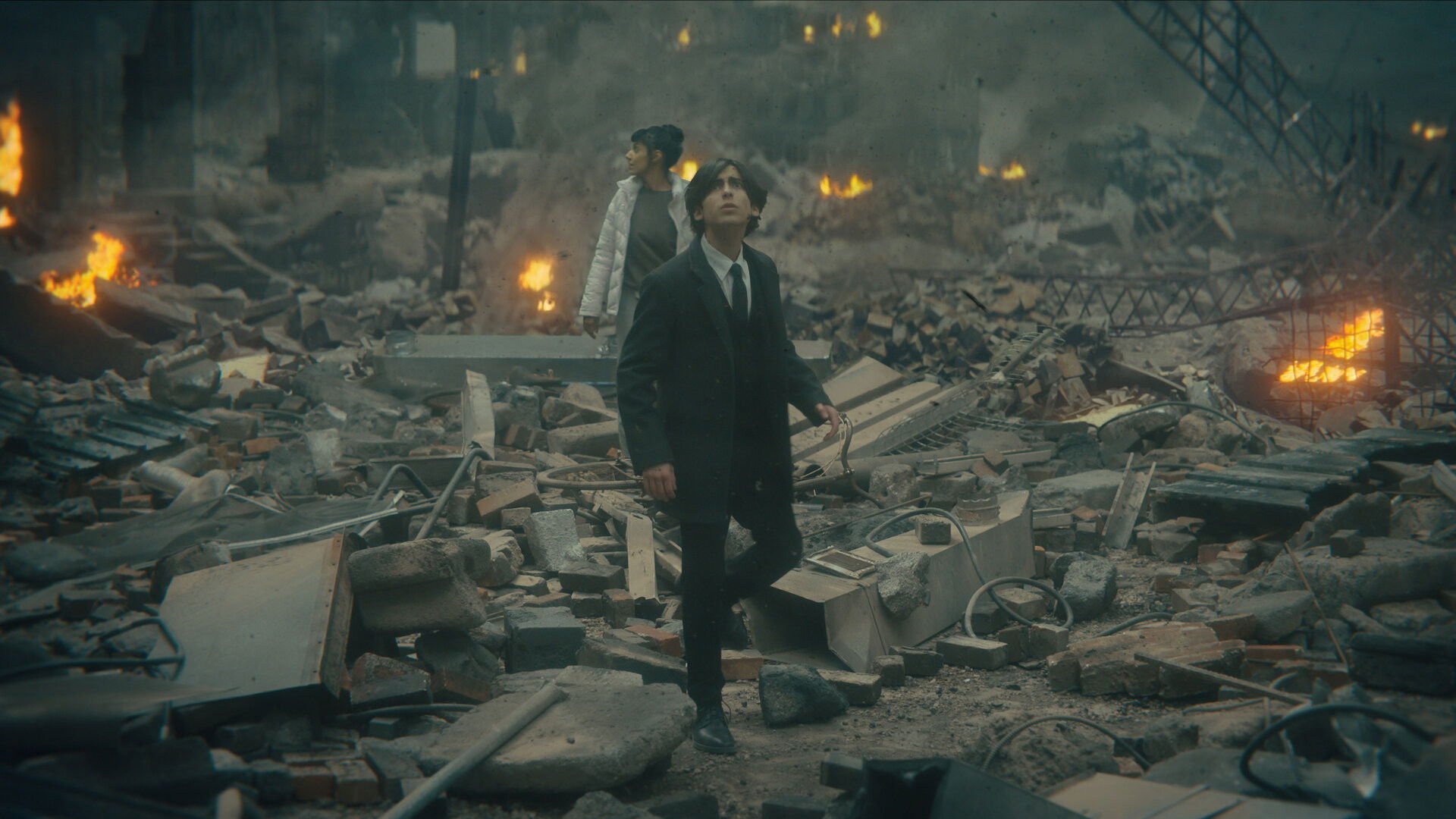The Umbrella Academy kadr | film.org.pl Kadr z serialu The Umbrella Academy: mężczyzna w ciemnym garniturze idzie przez zniszczone ruiny, w tle płoną ognie.