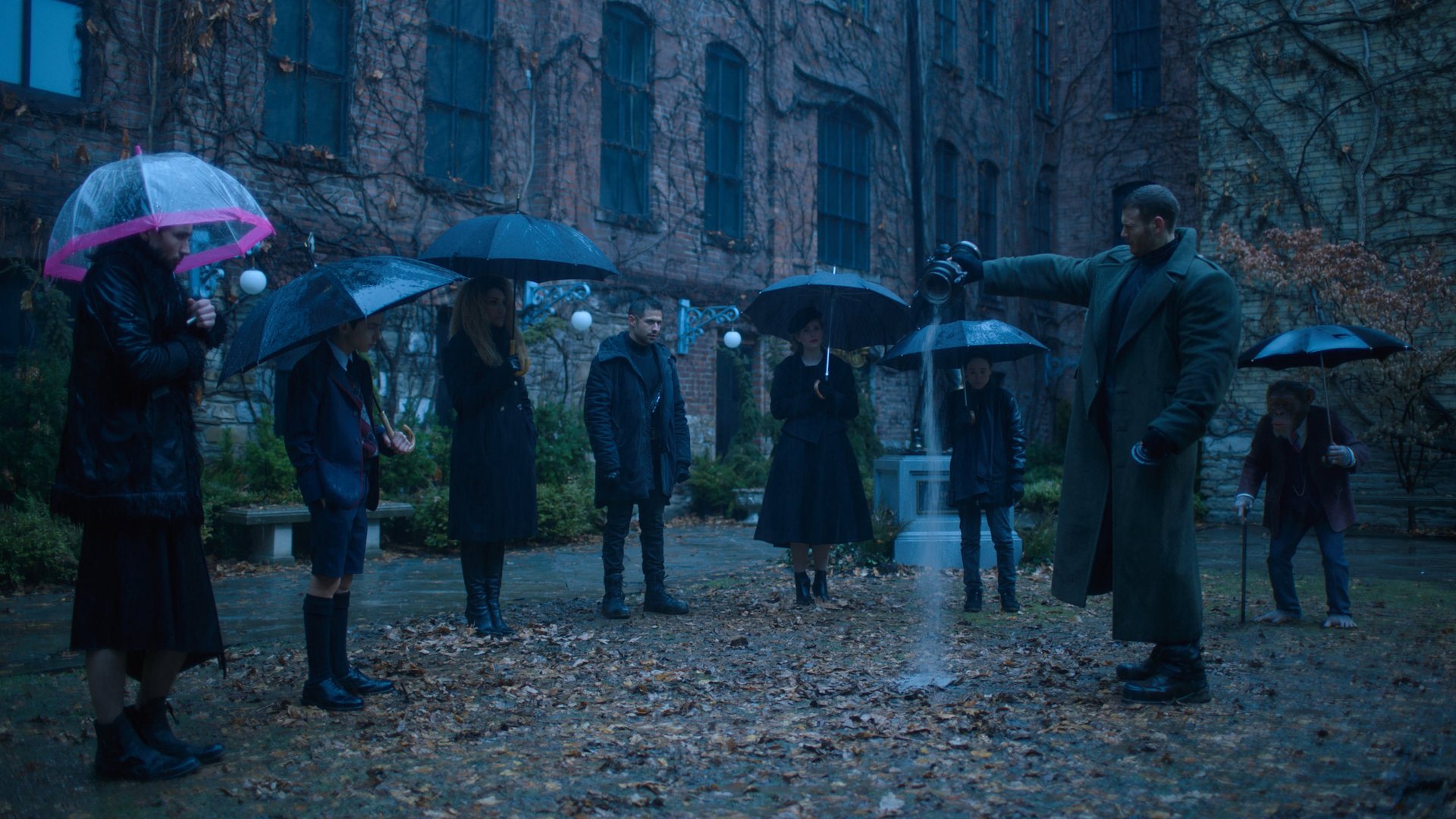 The Umbrella Academy kadr | film.org.pl grupa osób w ciemnych płaszczach stoi z parasolami na deszczowym dziedzińcu przy ceglanym budynku.