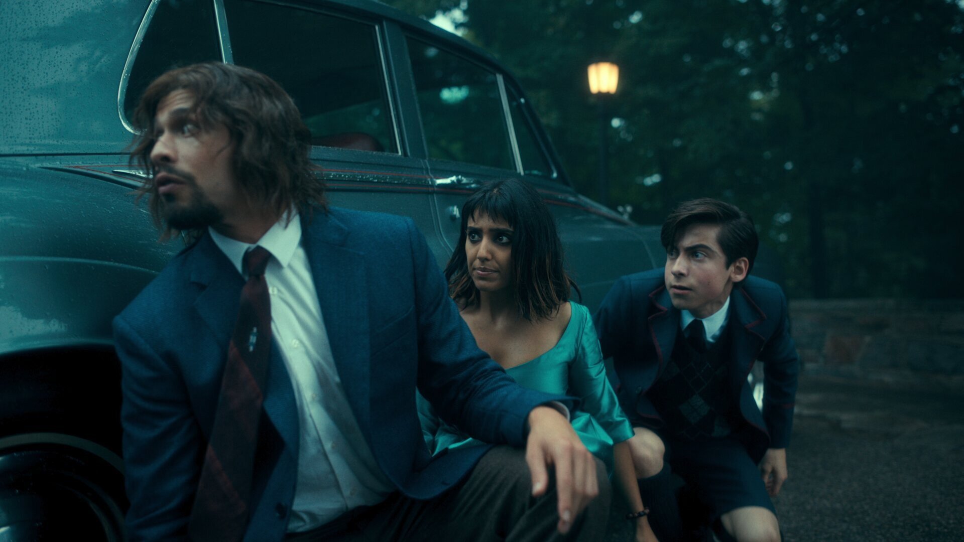 The Umbrella Academy kadr | film.org.pl Kadr z serialu The Umbrella Academy: trzy osoby w garniturach stoją przy ciemnozielonym samochodzie w nocnym parku, przy latarni.