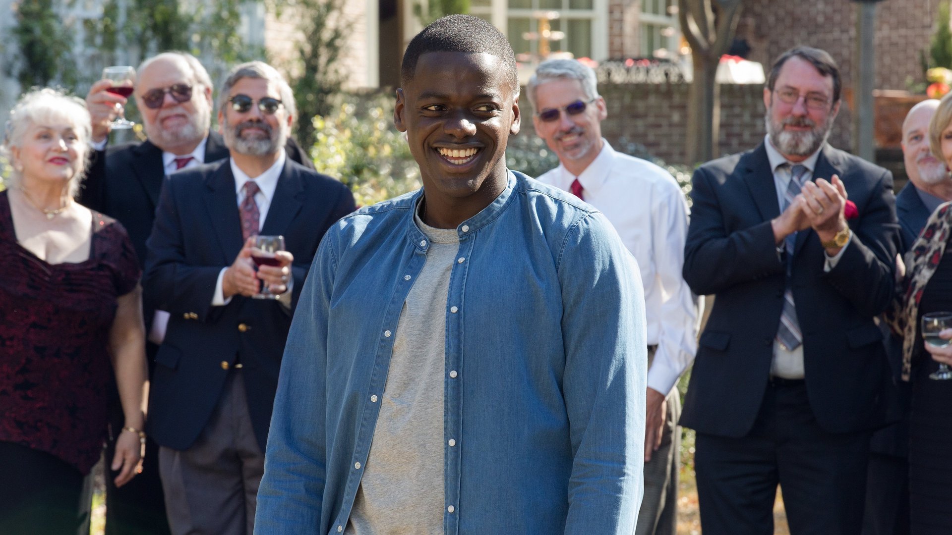 Uciekaj Get Out kadr | film.org.pl Kadr z filmu Uciekaj: uśmiechnięty młody mężczyzna w niebieskiej koszuli na tle gości w ogrodzie, z kieliszkami w dłoniach.