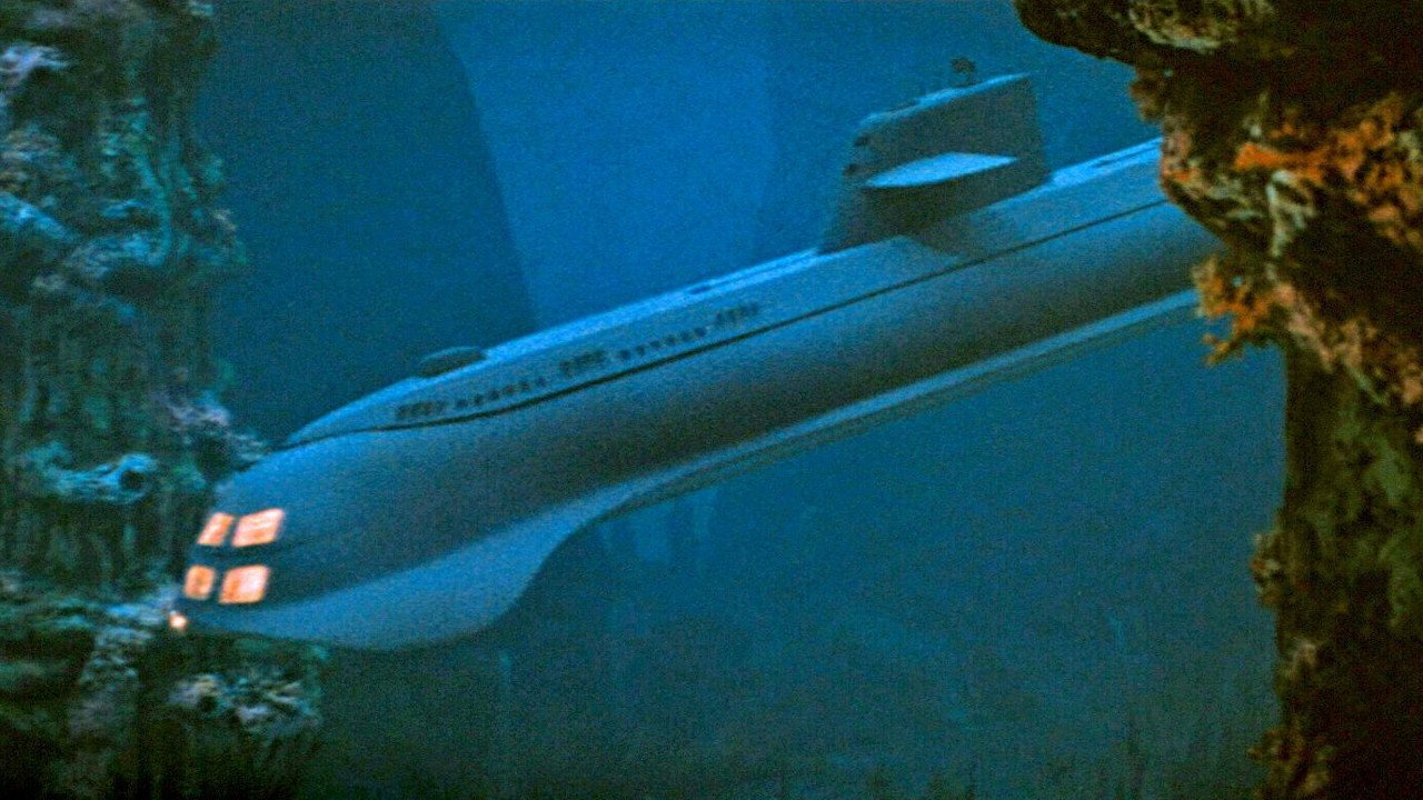 Kadr z filmu Voyage to the Bottom of the Sea: podwodna łódź podwodna z czerwonymi światłami przy dziobie, wśród skał i błękitnej wody.