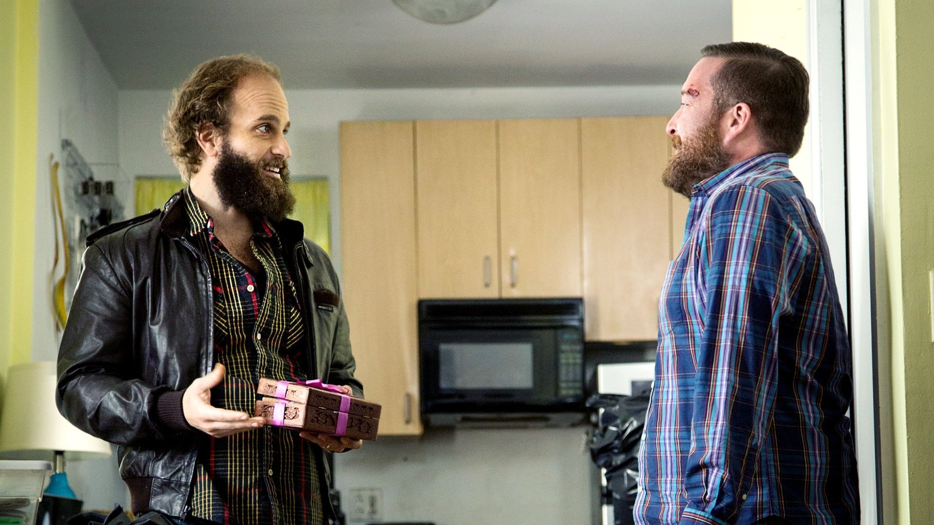 W potrzebie High Maintenance kadr | film.org.pl Kadr z serialu W potrzebie: dwaj brodaci mężczyźni w kuchni, jeden w skórzanej kurtce trzyma małe pudełko w kolorowe paski, drugi w kraciastej koszuli patrzy na niego.