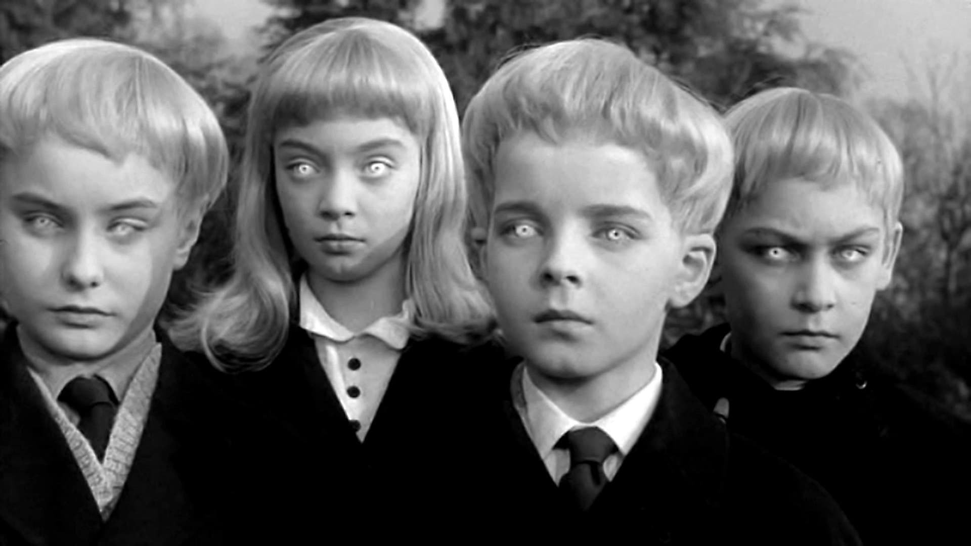 Kadr z filmu Wioska przeklętych: czworo jasnowłosych dzieci w ciemnych ubraniach patrzy w kamerę jasnymi, niemal białymi oczami.