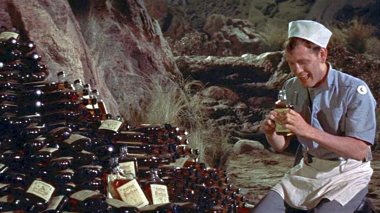 Zakazana planeta Forbidden Planet kadr | film.org.pl Kadr z filmu Zakazana planeta: mężczyzna w białej czapce siedzi obok ciemnych butelek z etykietami na skalistym terenie. space opera